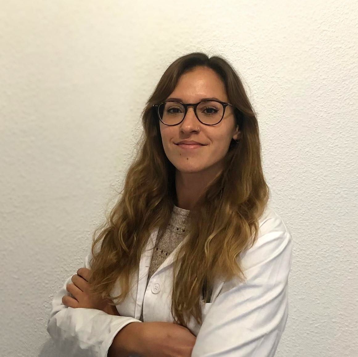 Una mujer que lleva gafas y una bata blanca está de pie con los brazos cruzados.
