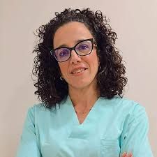 Una mujer que lleva una camisa blanca y un collar está parada frente a una pared blanca.