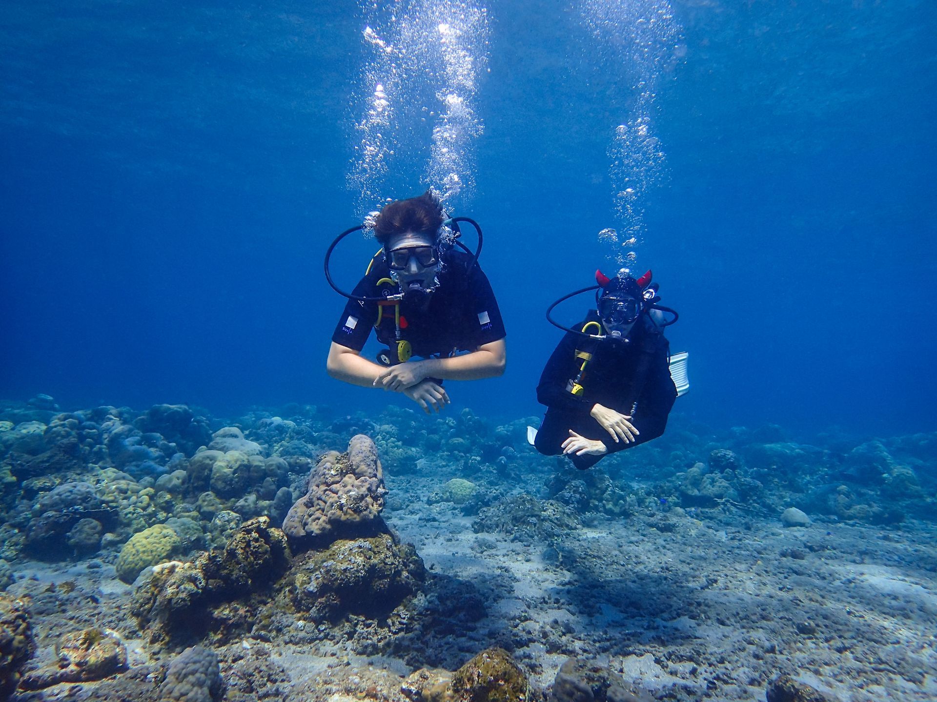Deux plongeurs sous-marins dans l'océan, des bulles remontent à la surface. Ils sont près d'un récif corallien, dans une eau bleue.