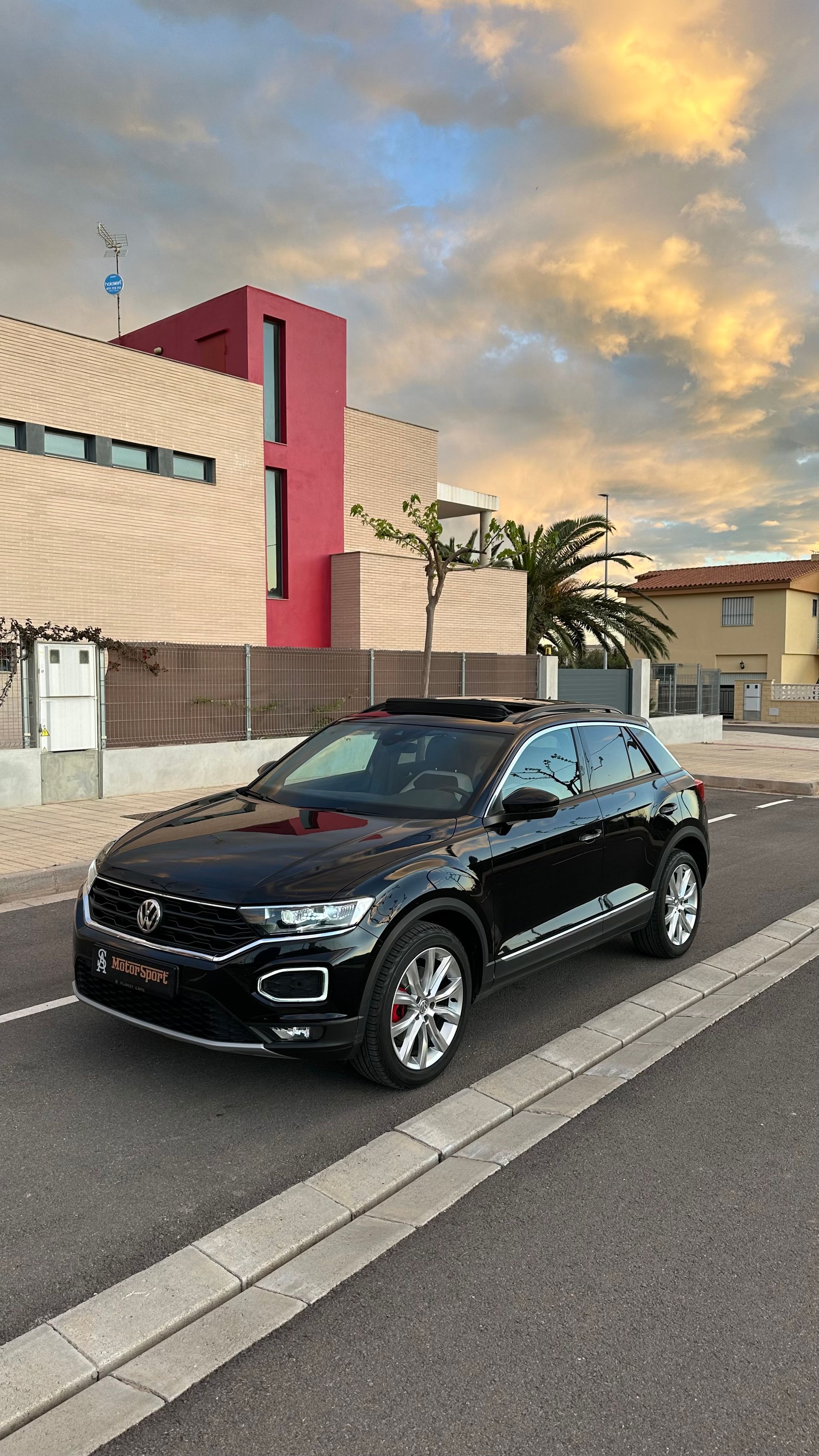 SUV Volkswagen negro estacionado en una calle con edificios y una puesta de sol al fondo.
