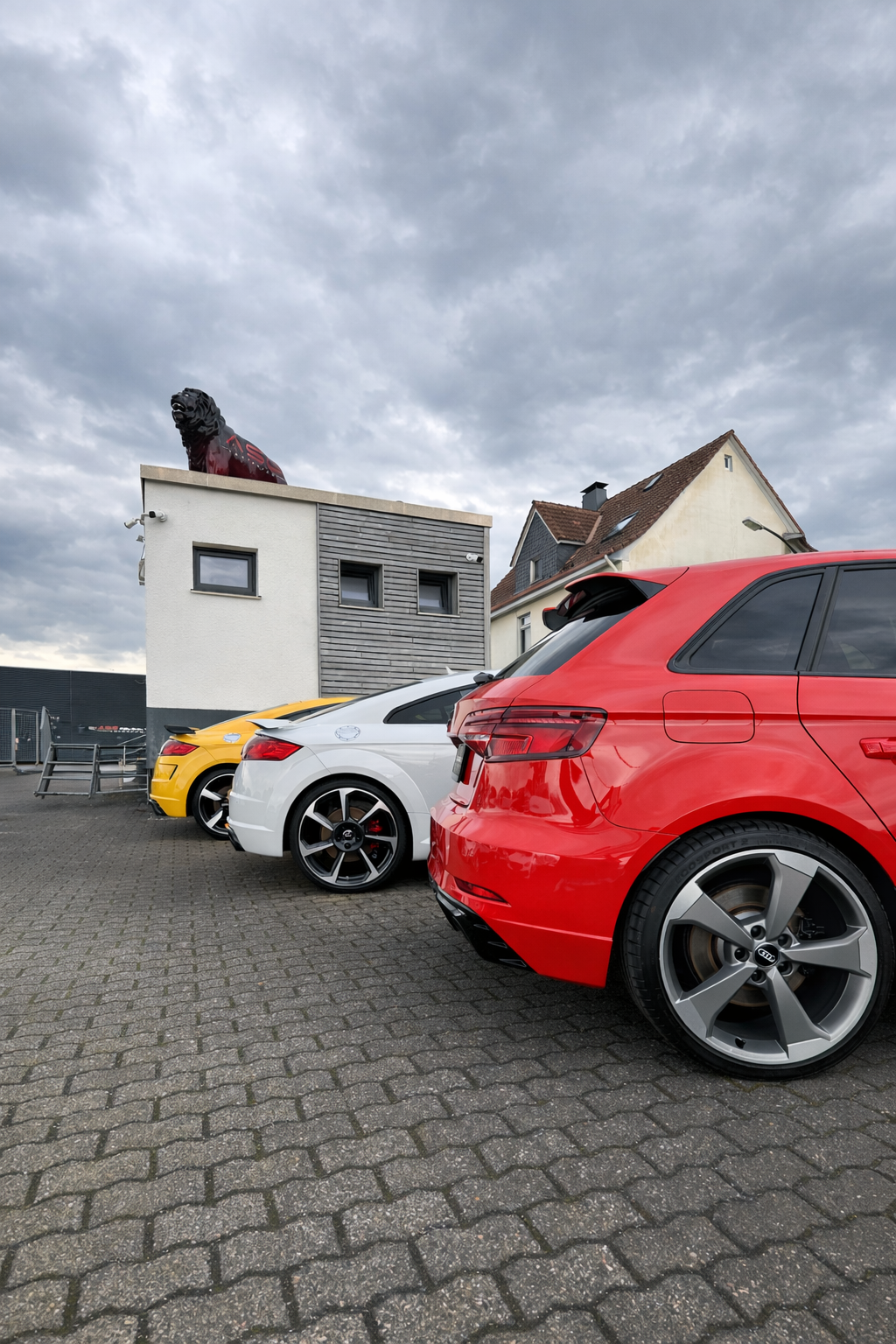 Tres coches: amarillo, blanco y rojo, estacionados sobre adoquines. Un edificio con una estatua de león y cielo nublado.