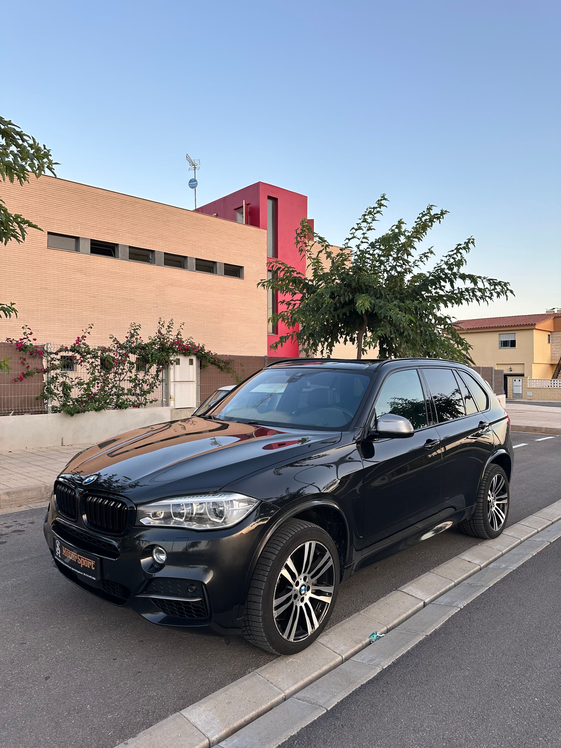 SUV BMW X5 negro estacionado en una calle frente a un edificio con detalles en rojo.