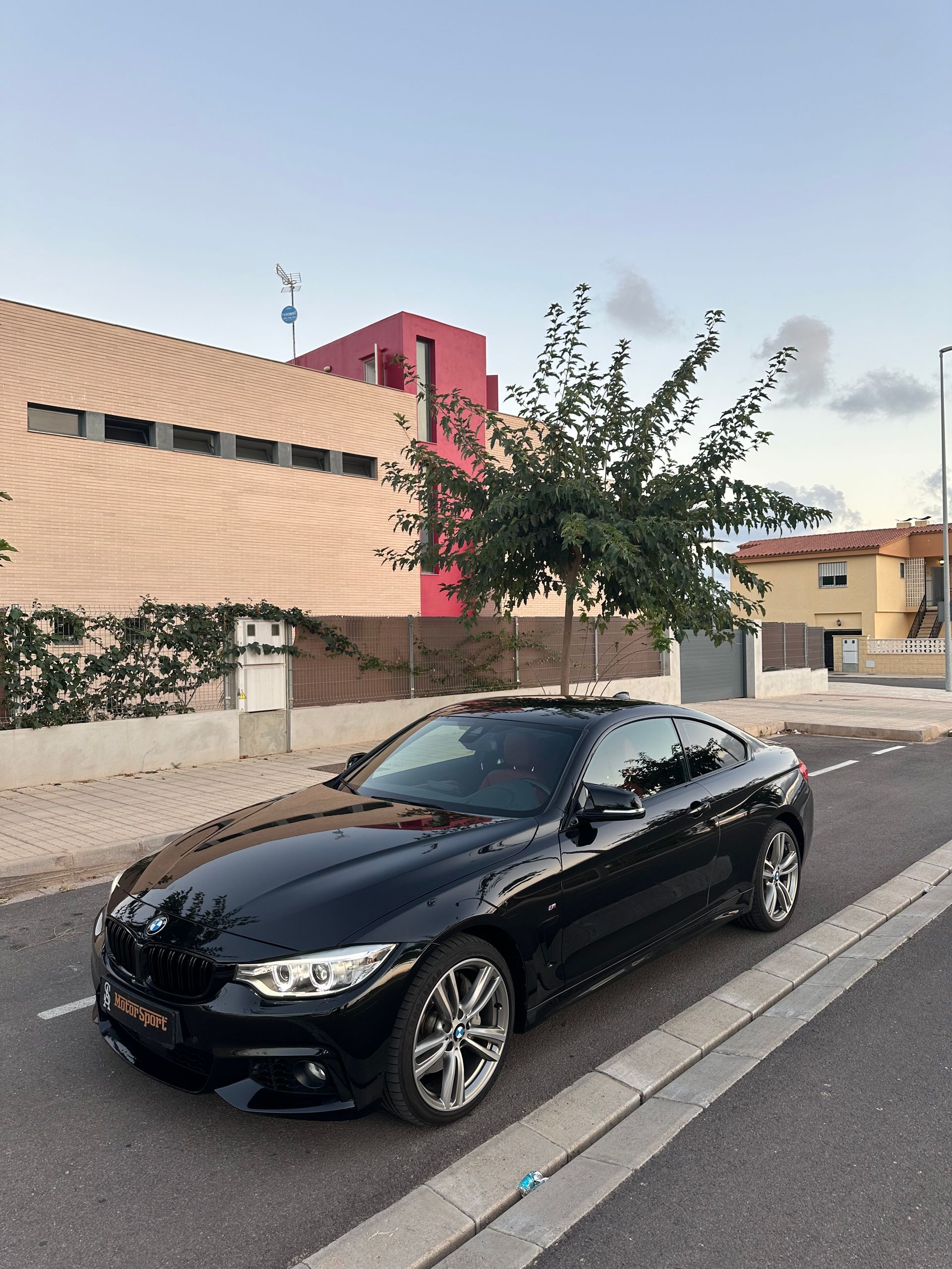BMW cupé negro estacionado en una calle con un edificio y un árbol al fondo.