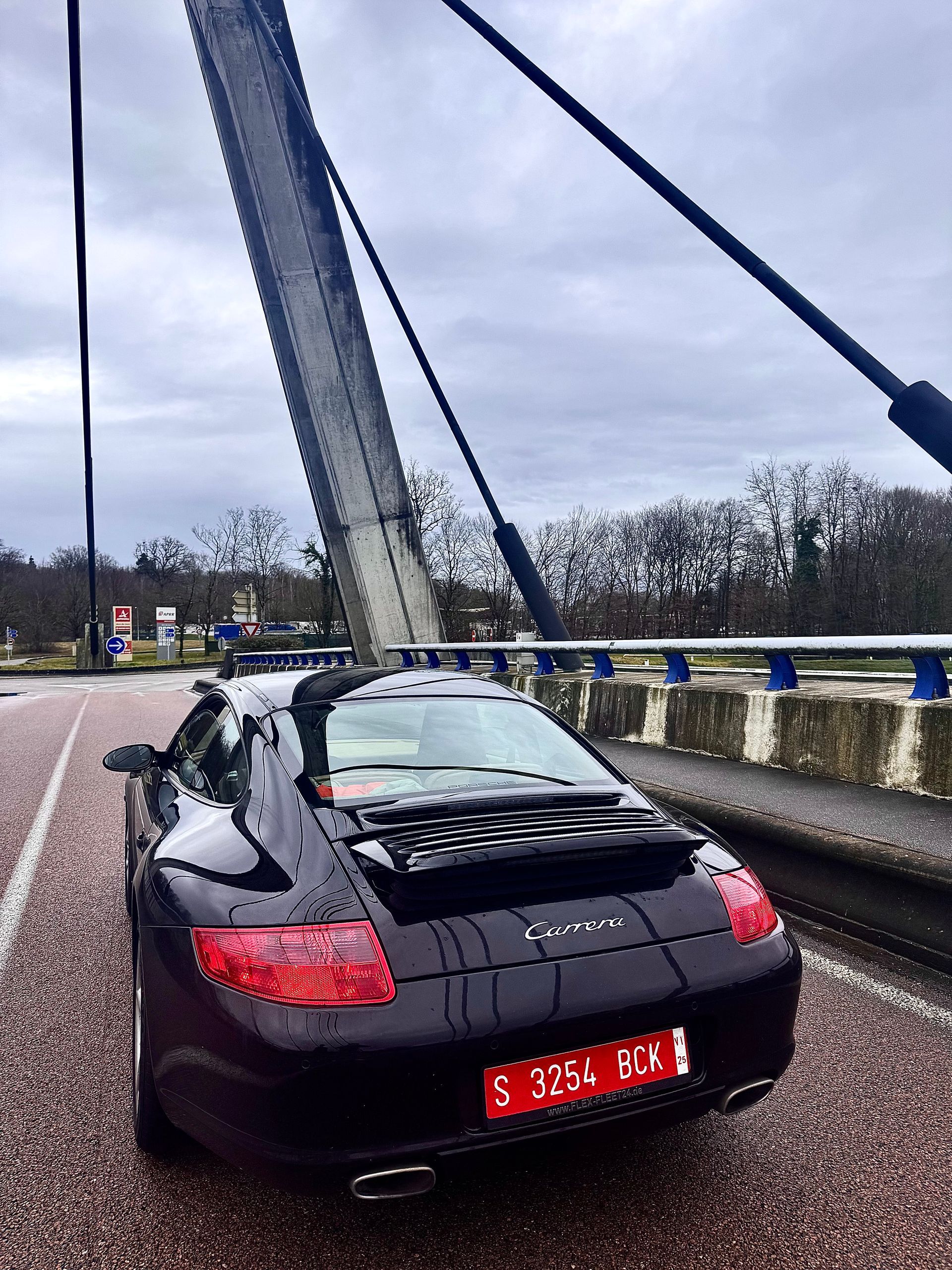 Porsche 911 Carrera negro sobre un puente, matrícula europea, cielo nublado.