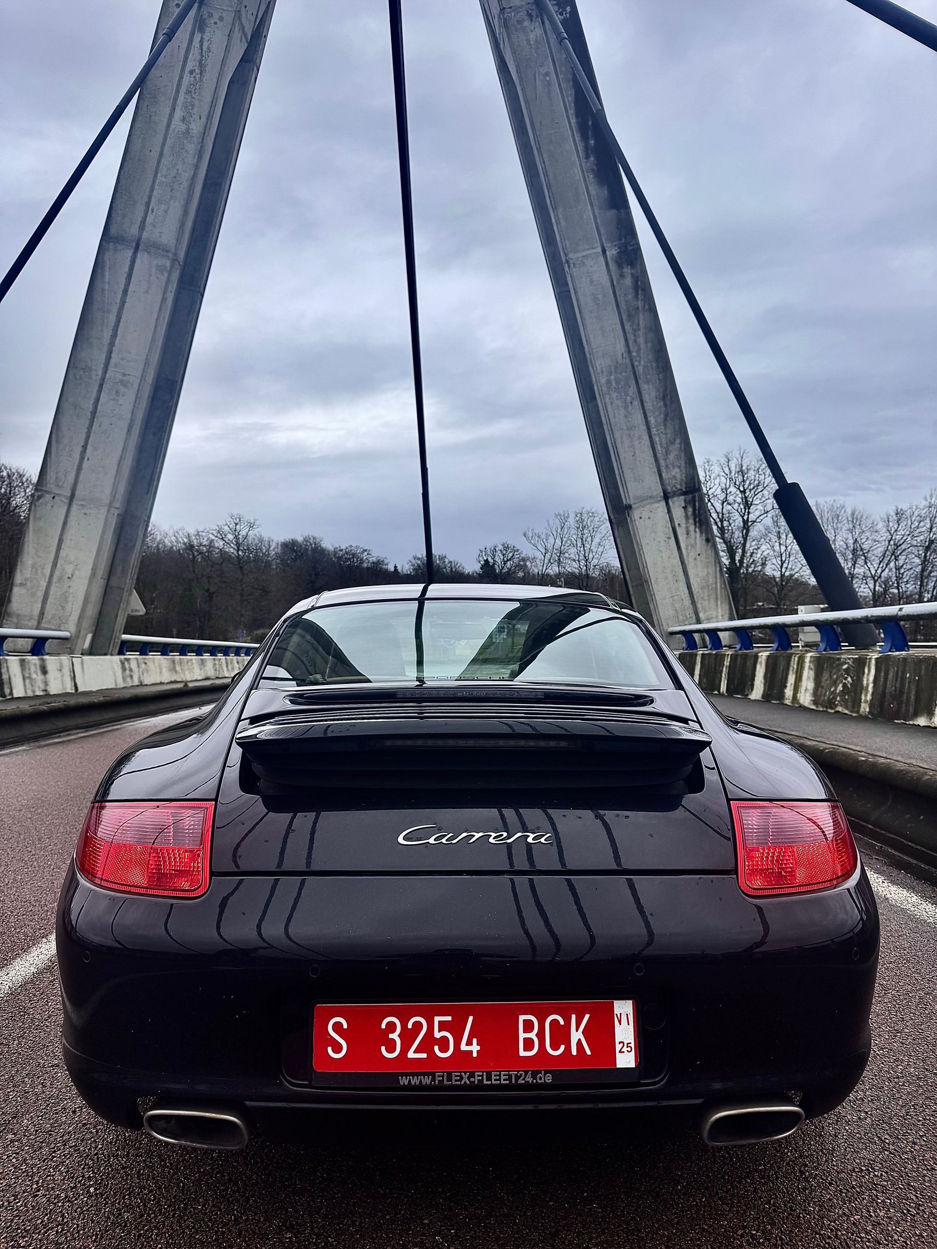 Porsche Carrera negro sobre un puente, vista trasera. La matrícula roja dice