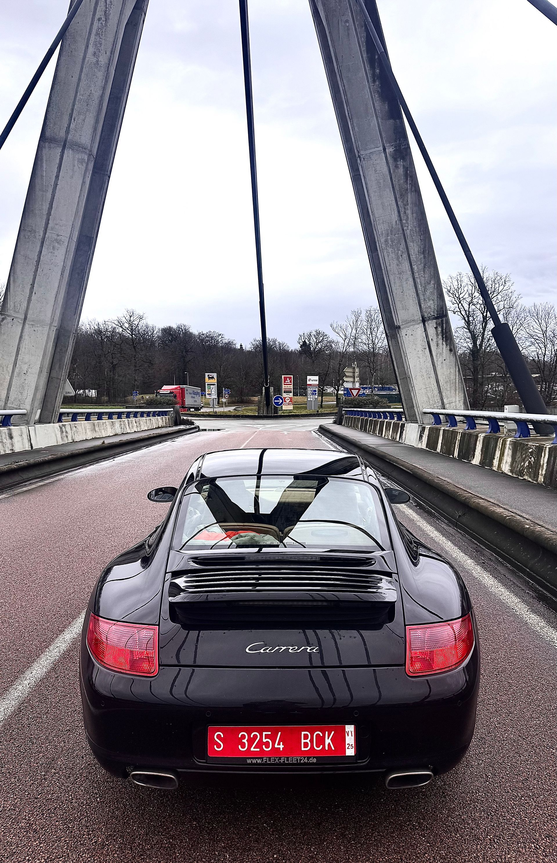 Porsche Carrera negro sobre un puente, vista trasera, bajo un cielo nublado.
