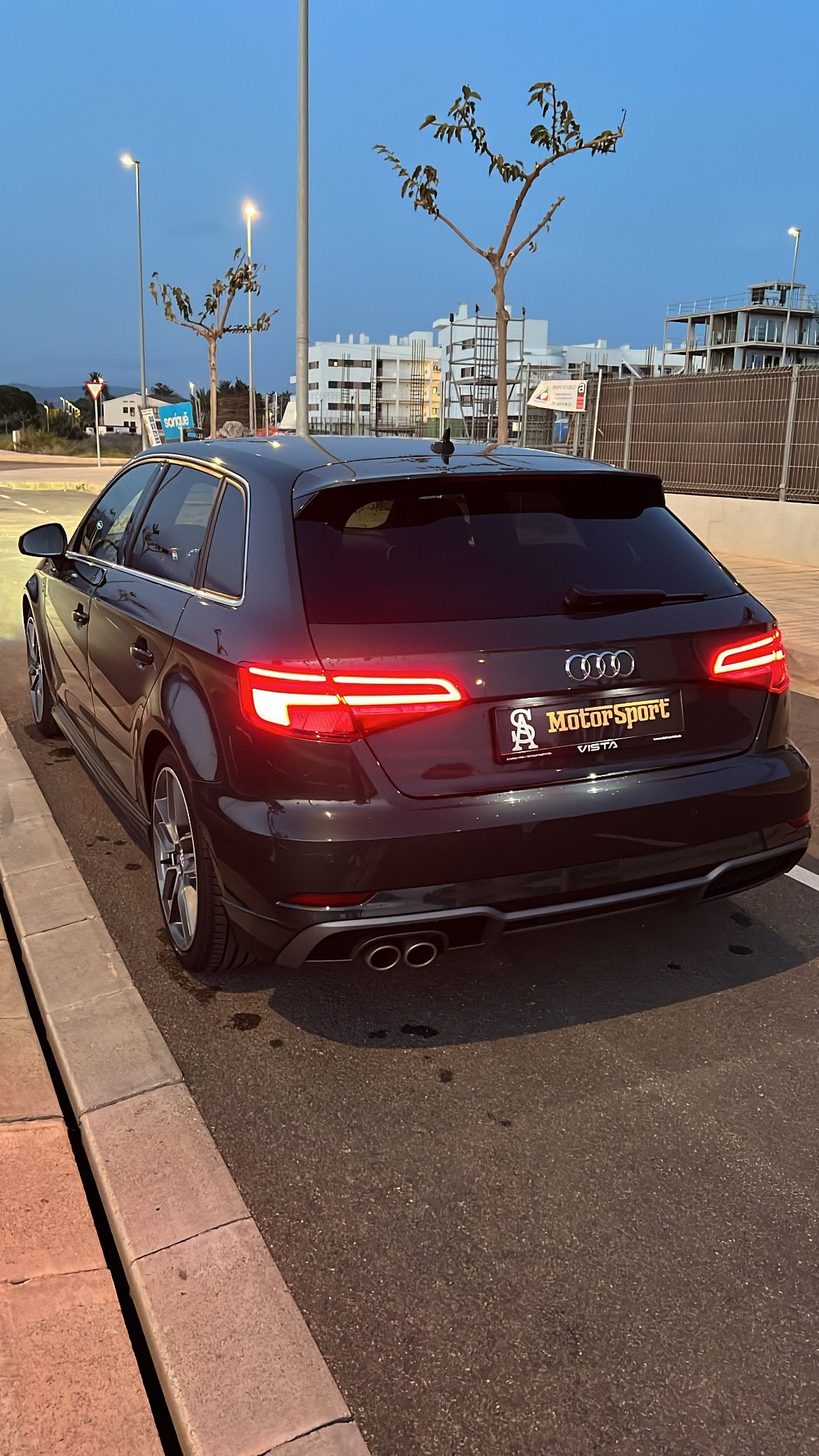 Audi hatchback negro estacionado al costado de una carretera, con las luces traseras iluminadas y un edificio de fondo.