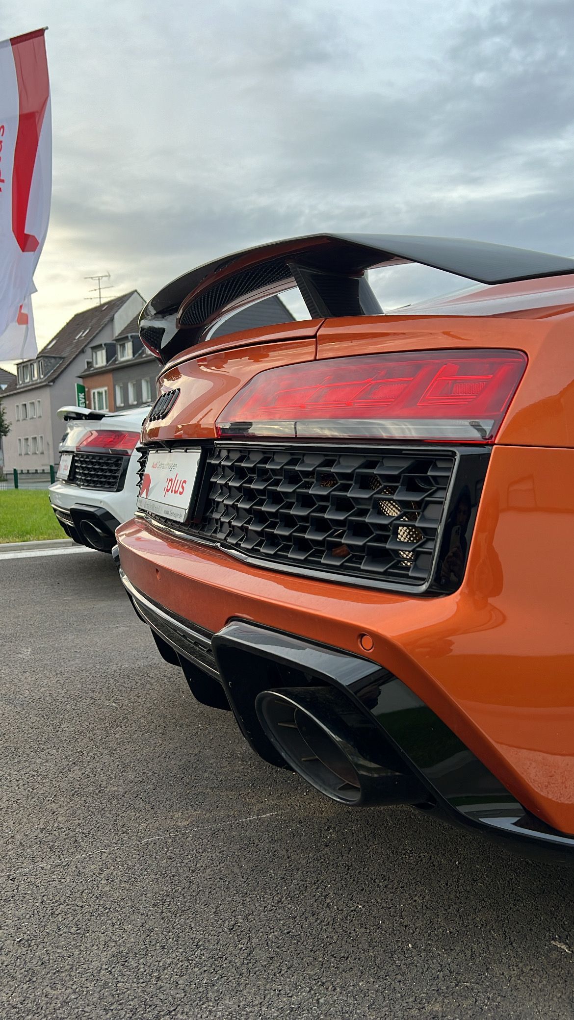 Audi R8 deportivo naranja, vista trasera, con detalles en negro. Otro coche visible al fondo.