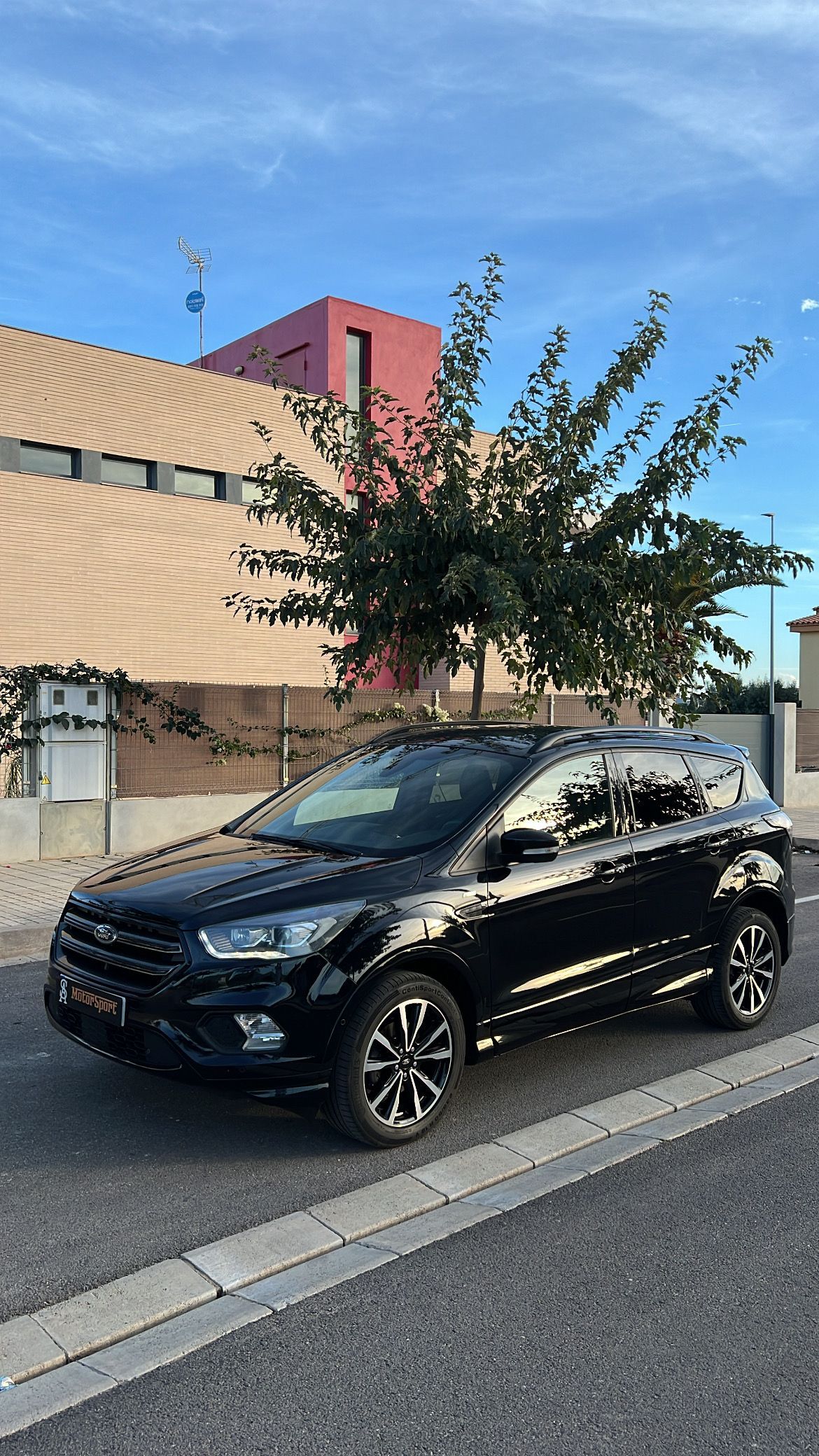 SUV negro estacionado en una calle frente a un edificio con paredes rojas y tostadas; árbol cercano bajo un cielo azul.