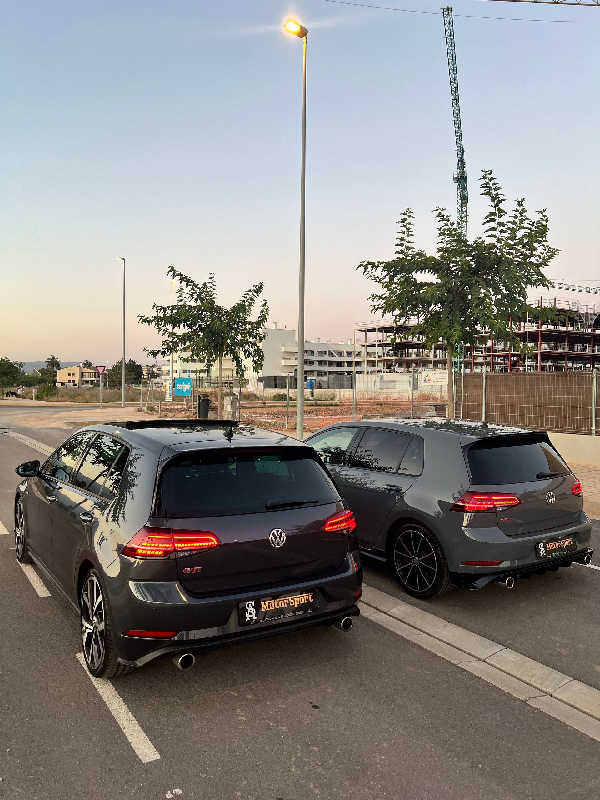 Dos coches Volkswagen Golf R color gris oscuro estacionados en una carretera con una farola, árboles y edificios al fondo.