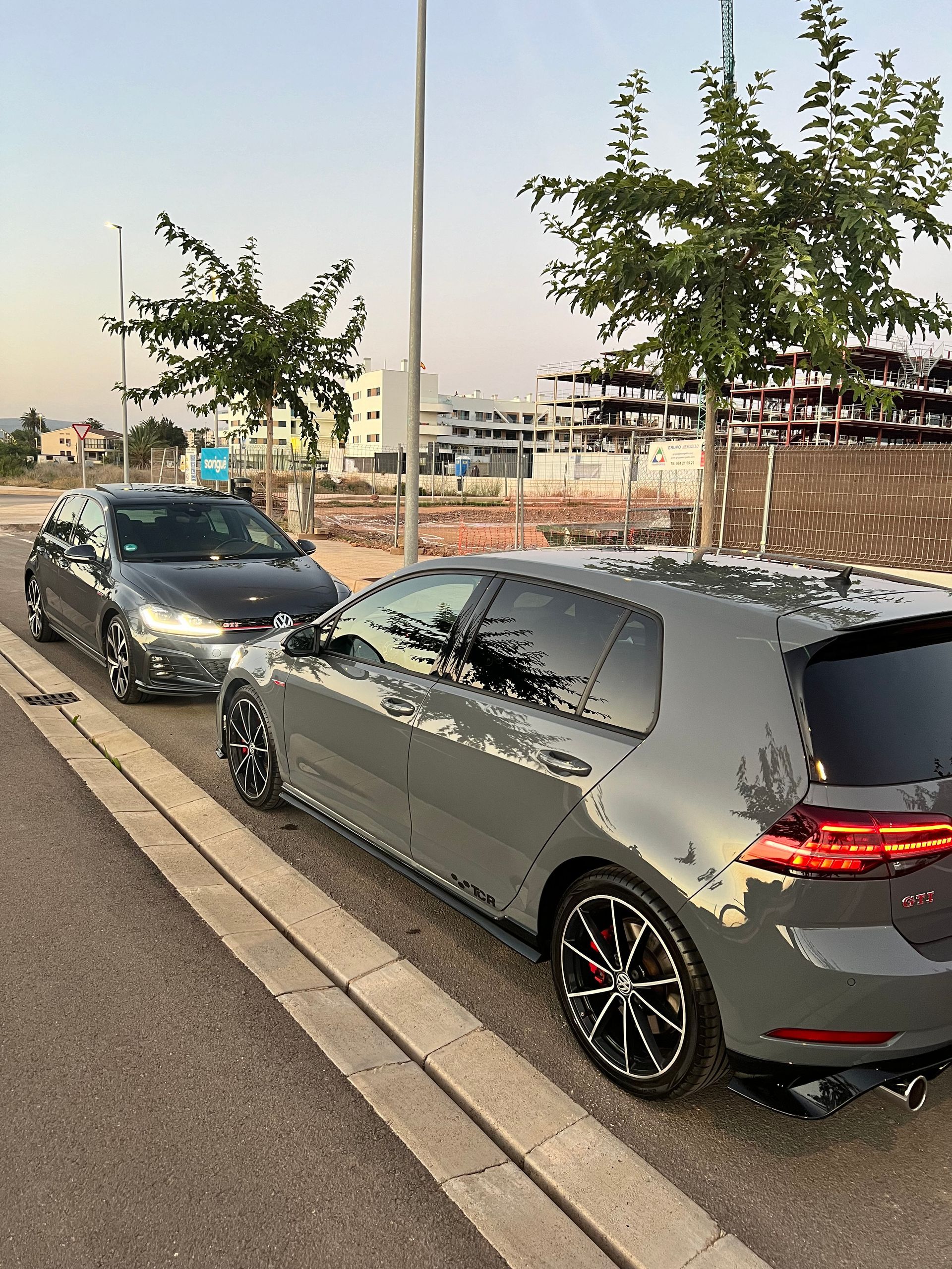 Dos hatchbacks grises estacionados a lo largo de una carretera con árboles y edificios al fondo.