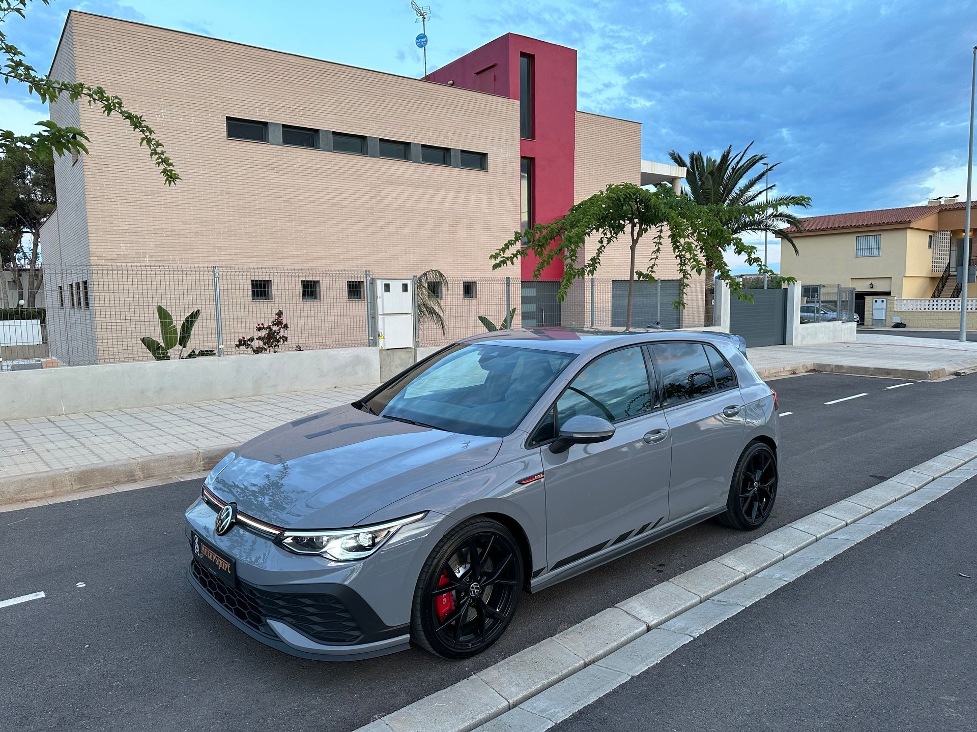 Volkswagen Golf GTI gris estacionado en una calle frente a un edificio moderno con detalles en rojo.