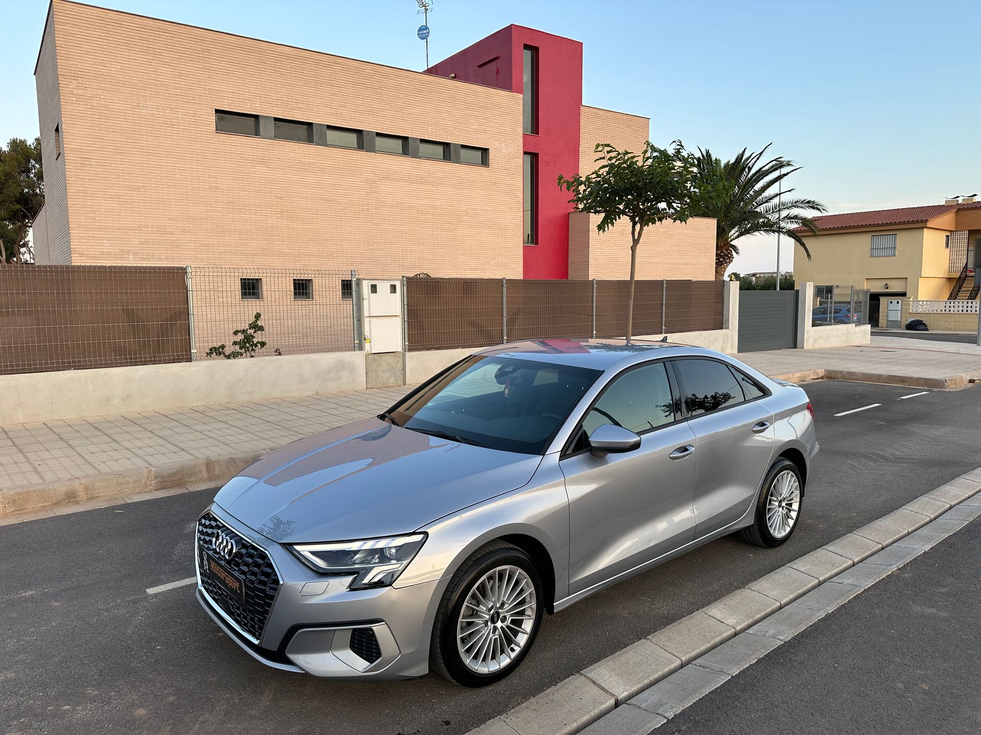 Un Audi sedán plateado estacionado en una calle frente a un moderno edificio de color beige y rojo.