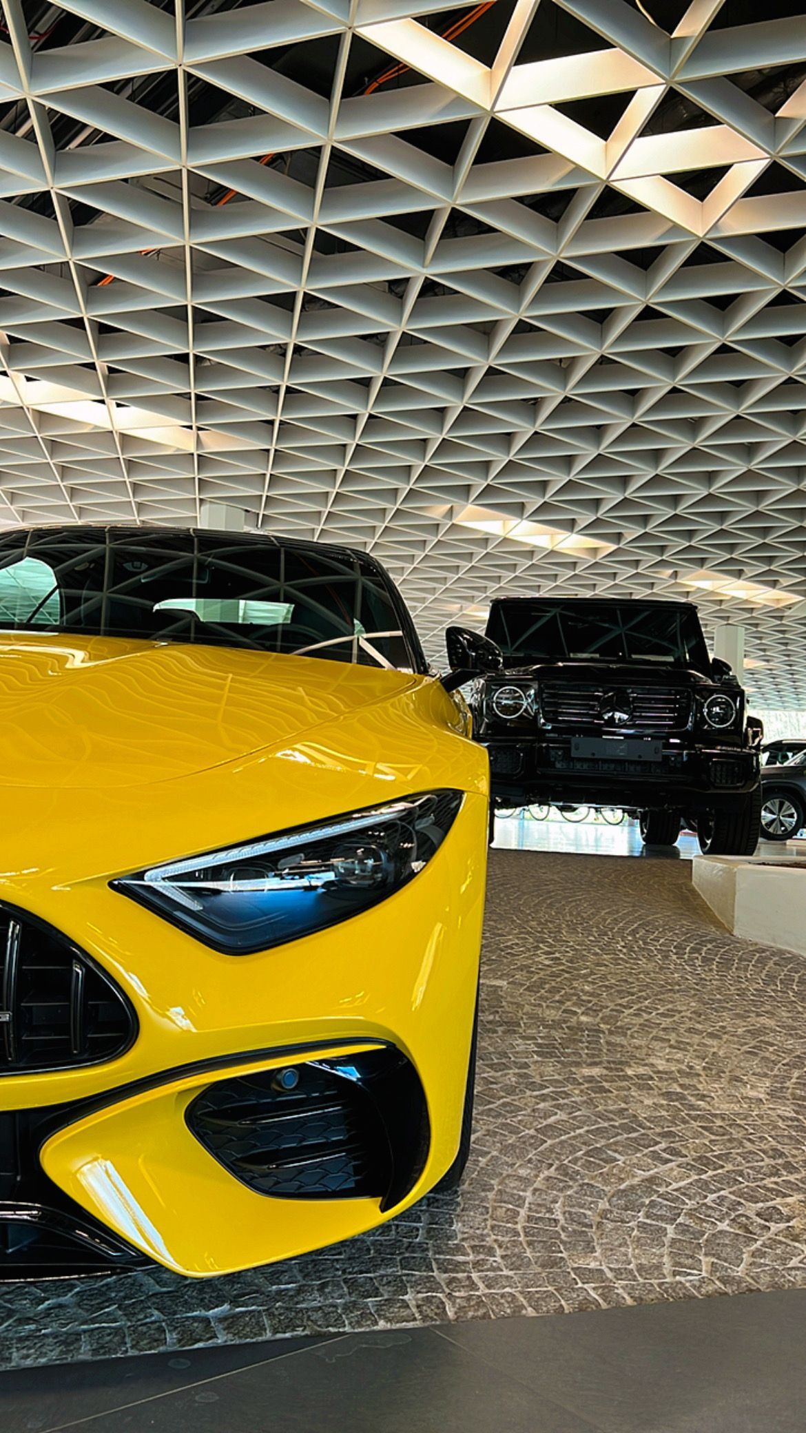 Coche deportivo amarillo en una sala de exposición con un vehículo negro al fondo, bajo un techo geométrico.