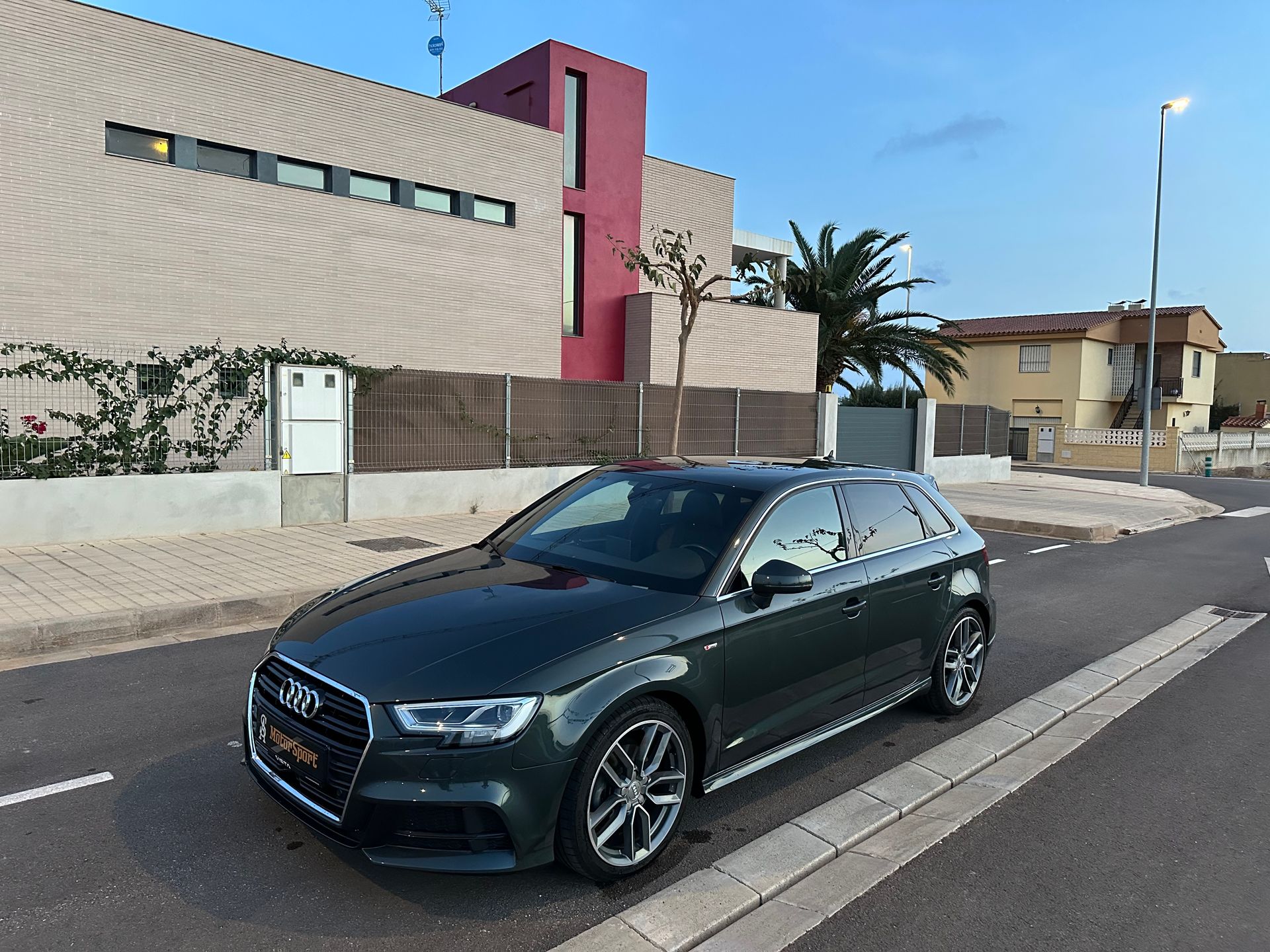 Audi A3 verde oscuro estacionado en una calle frente a un edificio moderno con detalles en rojo.