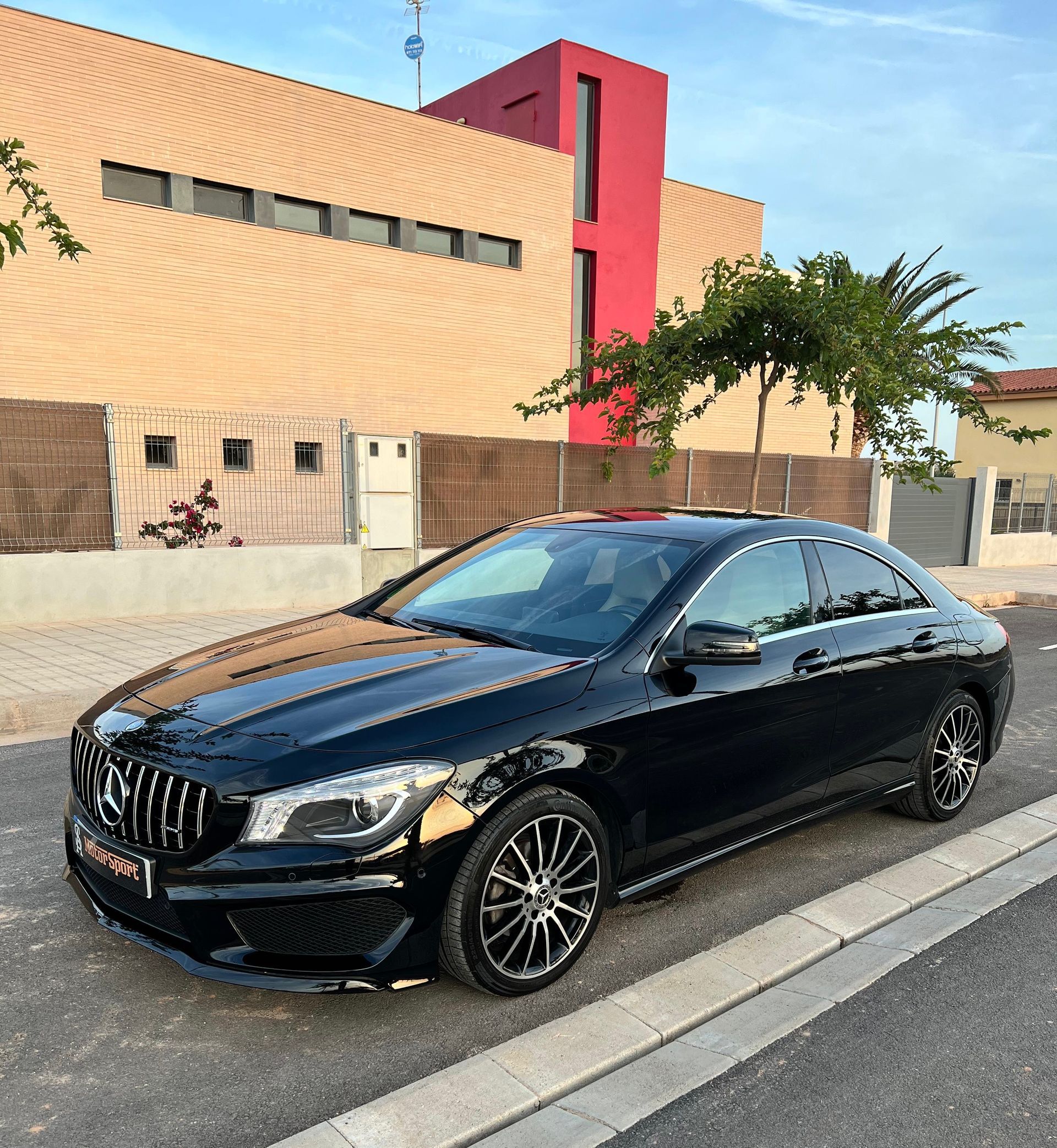 Un Mercedes-Benz CLA negro estacionado en una calle frente a un edificio de color tostado y rojo.