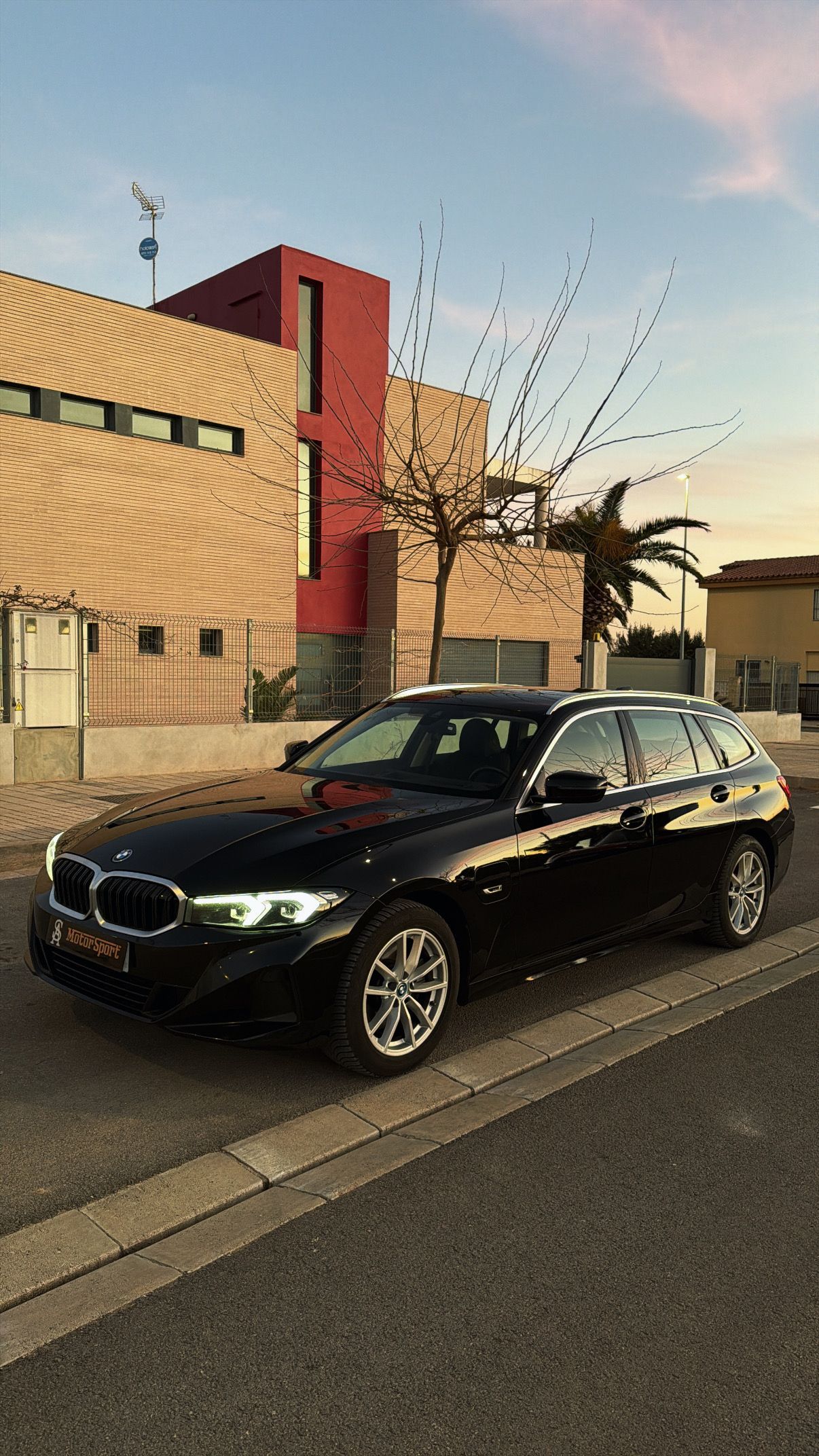 Una camioneta BMW negra estacionada en una calle frente a un edificio moderno; con la puesta de sol al fondo.