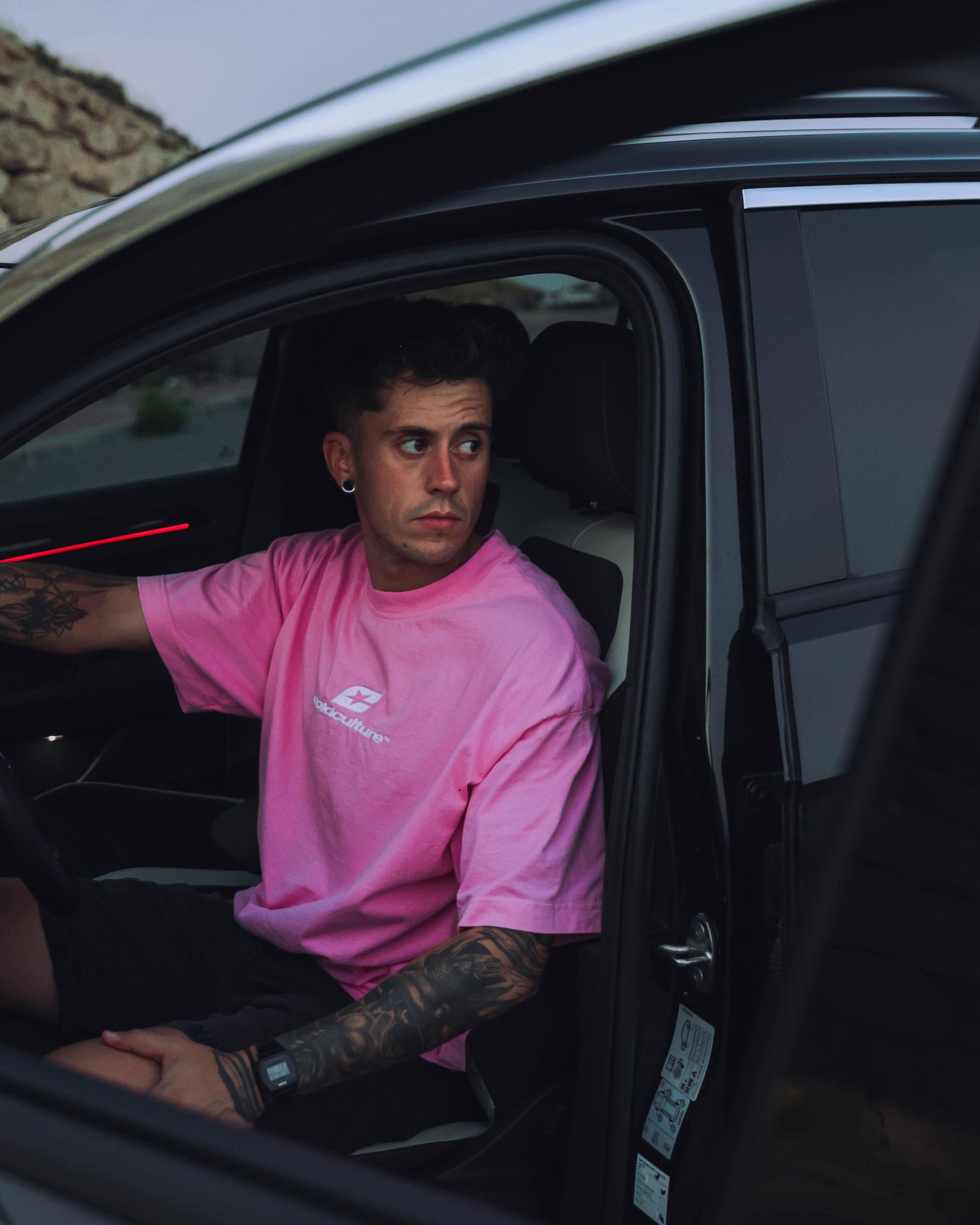 Un hombre con camisa rosa en un coche negro, mirando hacia un lado. Tiene tatuajes y sujeta el volante.