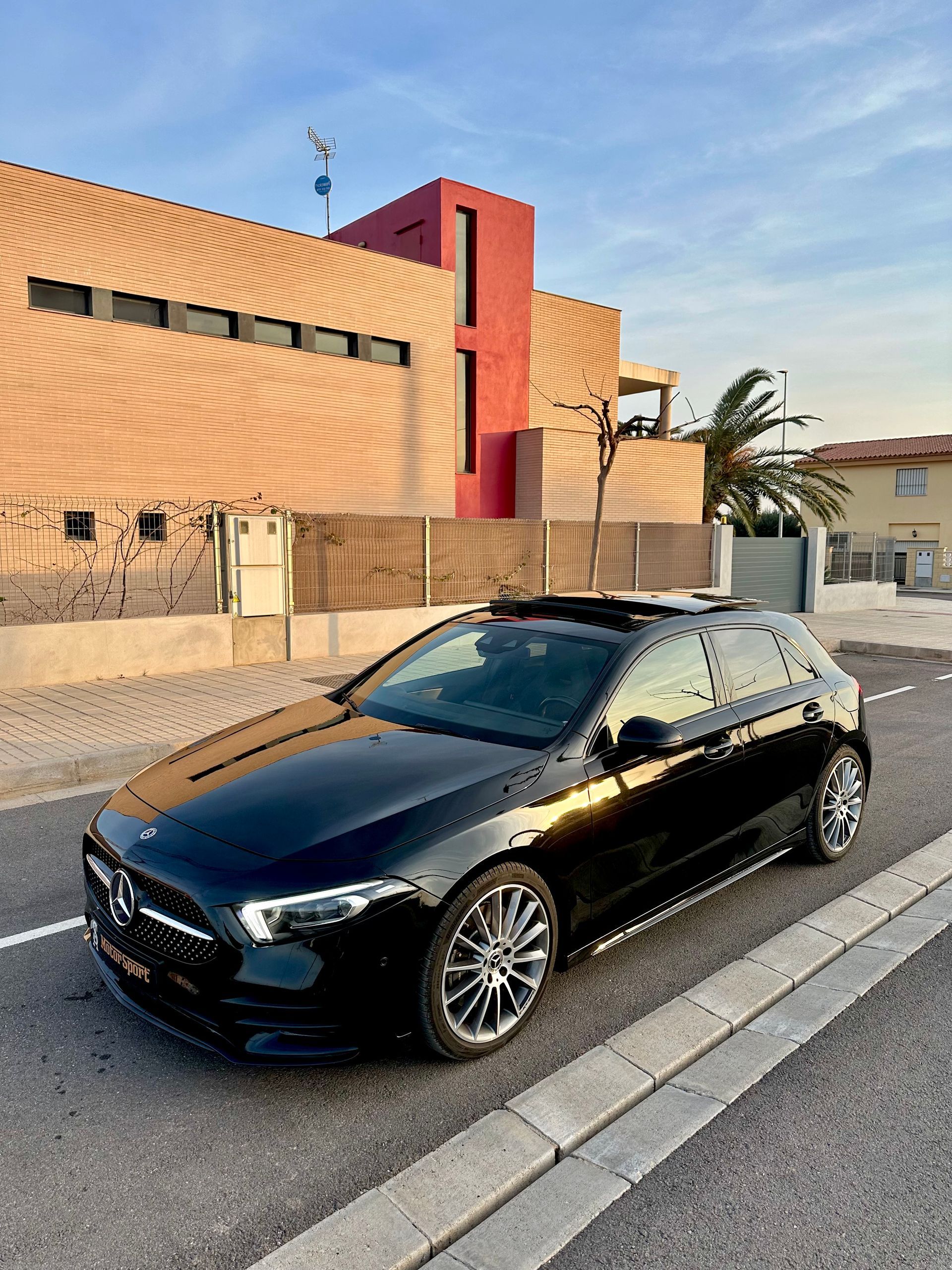 Un Mercedes-Benz hatchback negro estacionado en una calle frente a un edificio de color marrón rojizo en un día soleado.