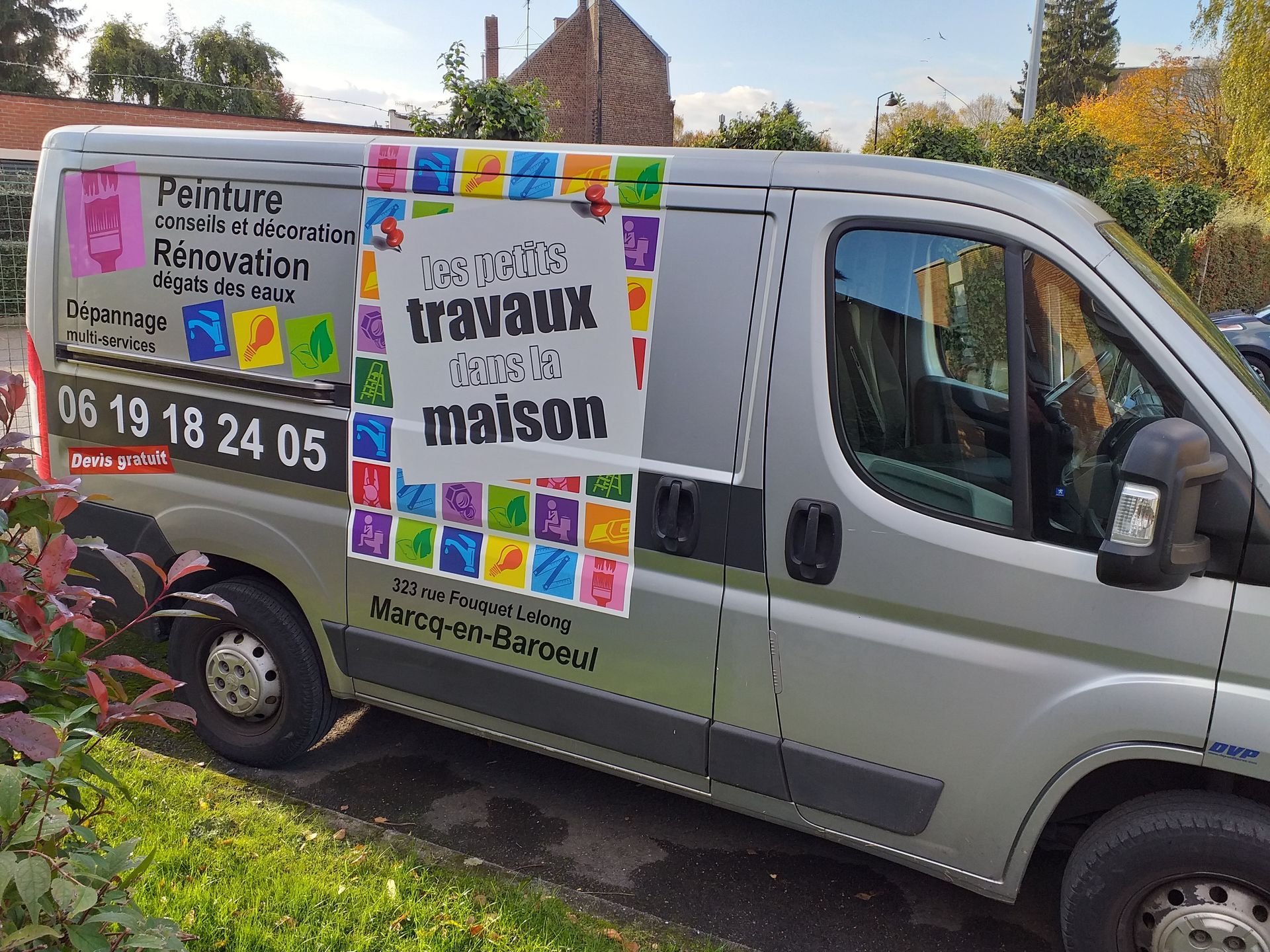 Fourgonnette argentée avec publicité des Petits Travaux Dans La Maison. Logo et numéro de téléphone colorés, garée dans la rue.