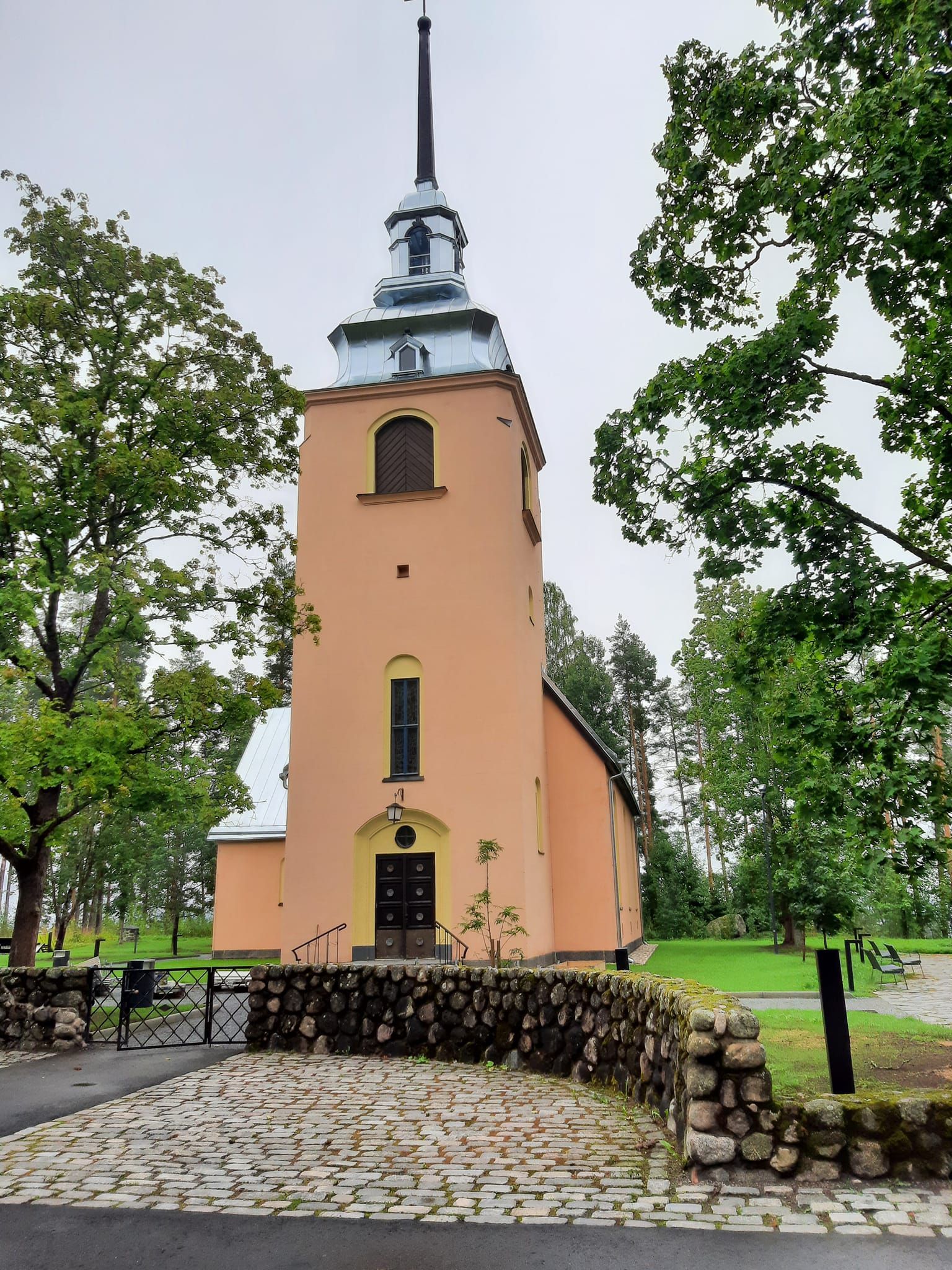 Vaaleanpunainen kirkko, jossa on korkea torni, kivimuuri ja puita pilvisessä, vehreässä ympäristössä.