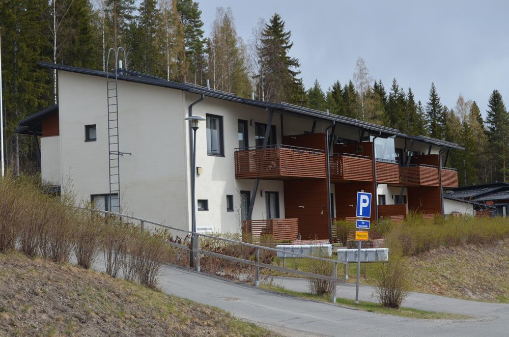 Moderni kaksikerroksinen kerrostalo, jossa on parvekkeet, tumma katto, vaaleat seinät ja pysäköintikyltti.