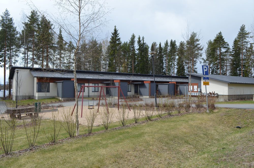 Leikkikenttä ja rakennuksia pysäköintialueella, puita taustalla.