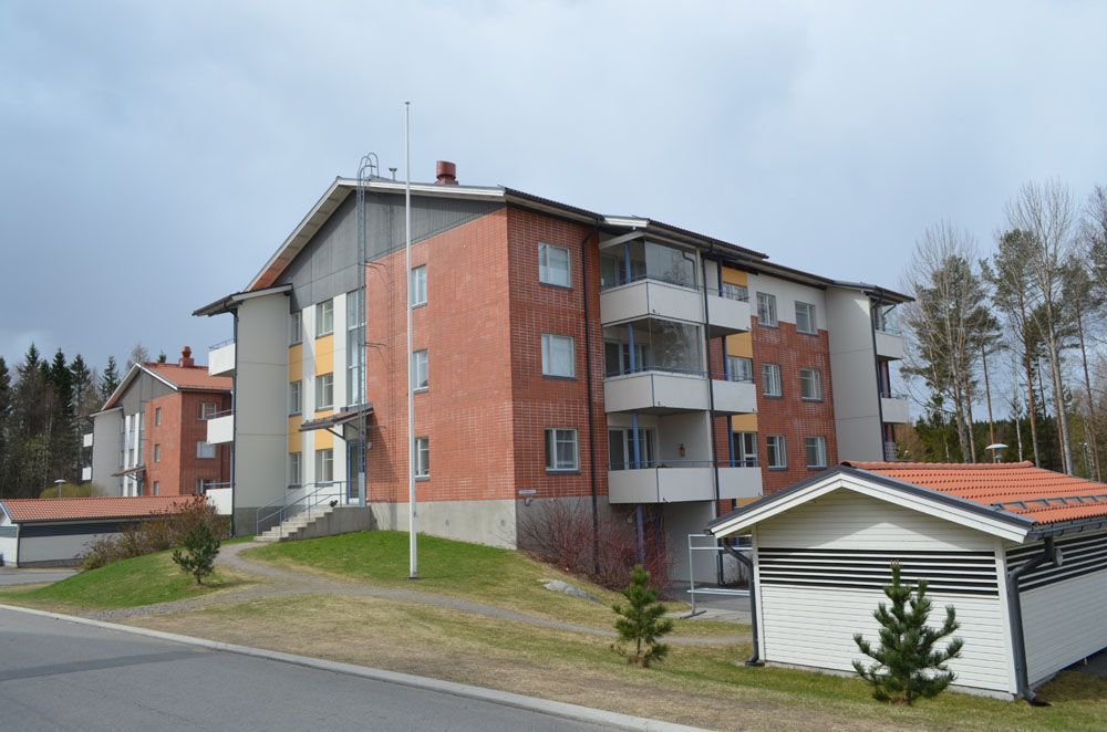 Punatiilinen kerrostalo, jossa on parvekkeita ja pieni ulkorakennus, loivalla kukkulalla pilvisen taivaan alla.