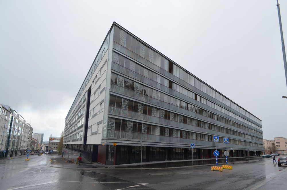 Moderni, kulmikas rakennus kaupungin risteyksessä pilvisenä päivänä; useita kerroksia, lasi-ikkunat.