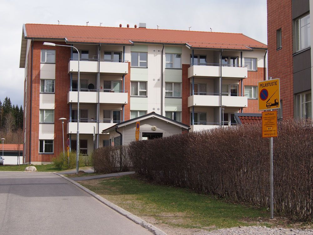 Parvekkeellinen ja punaisella tiilikatolla varustettu kerrostalo pensaikon ja asfalttitien vieressä.