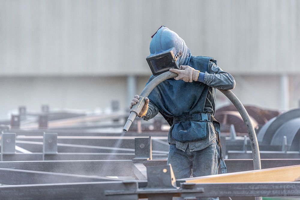 Eine Person sandstrahlt im Freien ein Metallgerüst; sie trägt Schutzausrüstung.