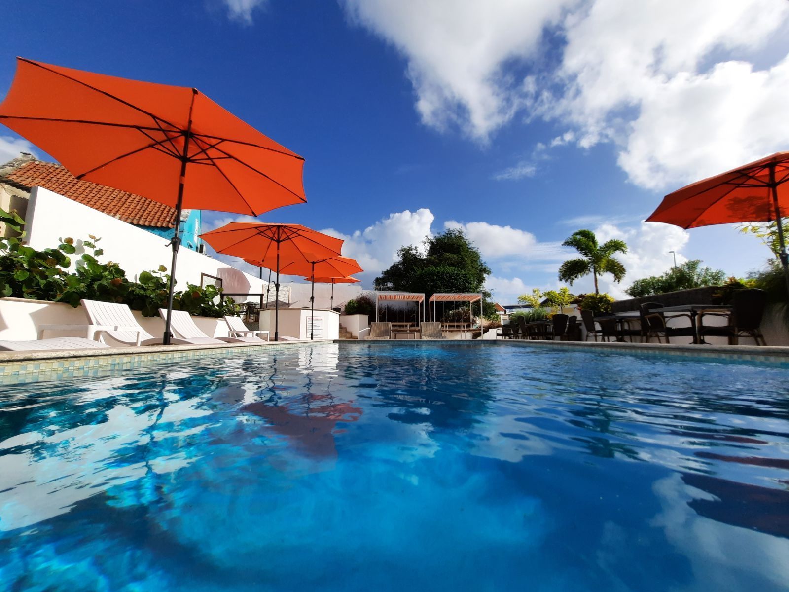 Poolside resort with blue water, orange umbrellas, lounge chairs, and a bright sky
