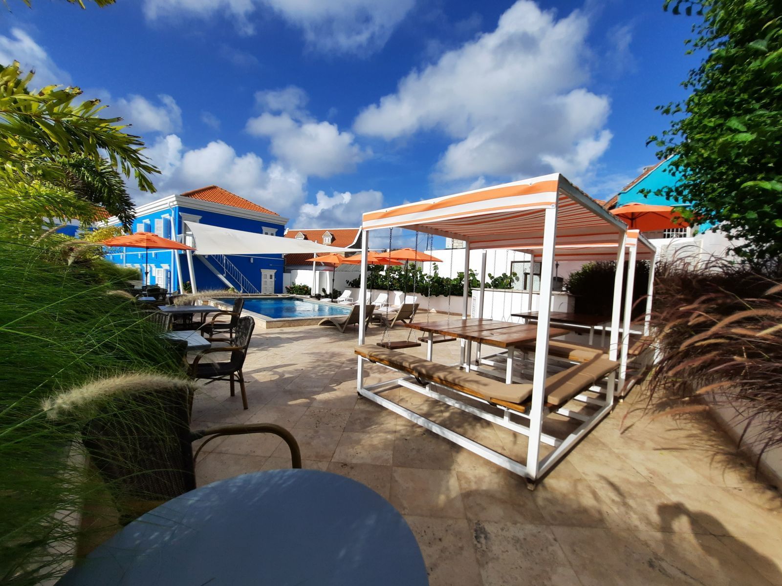 Sunny outdoor patio with white-framed loungers, orange canopies, and blue buildings under a bright sky