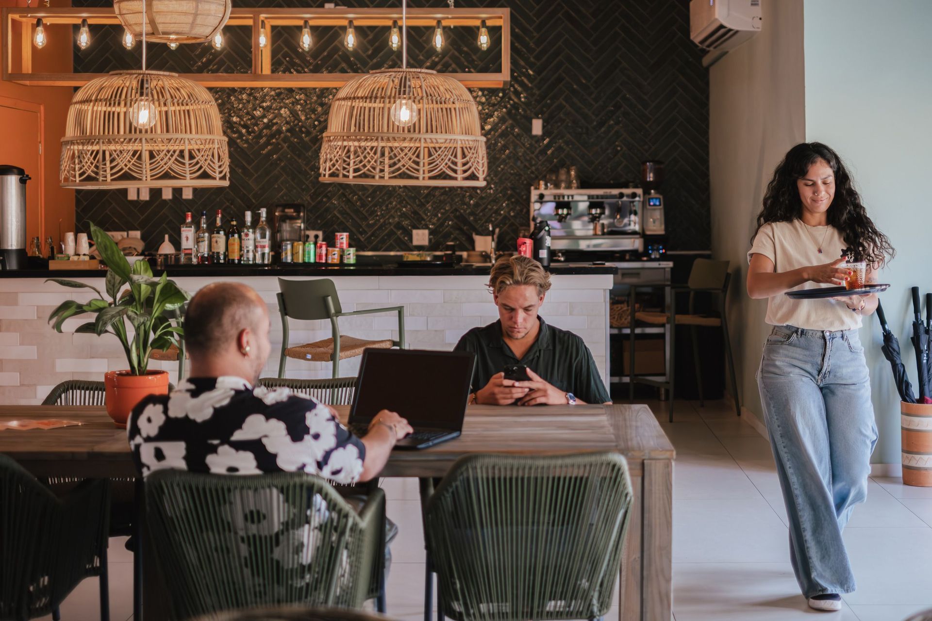 People seated and working in a cozy cafe, with warm pendant lights and a woman walking by carrying a tray.