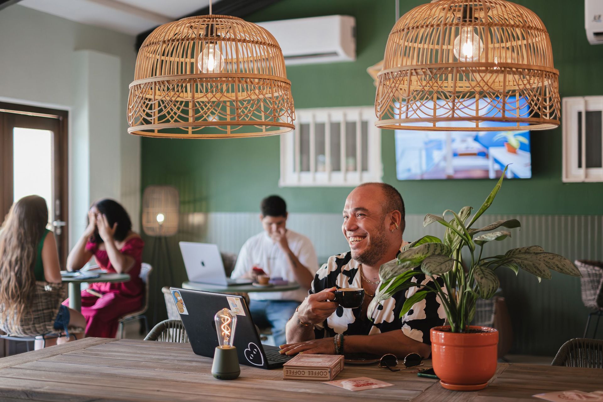 Café with people working and chatting, warm wicker pendant lights, man smiling at a laptop in the foreground