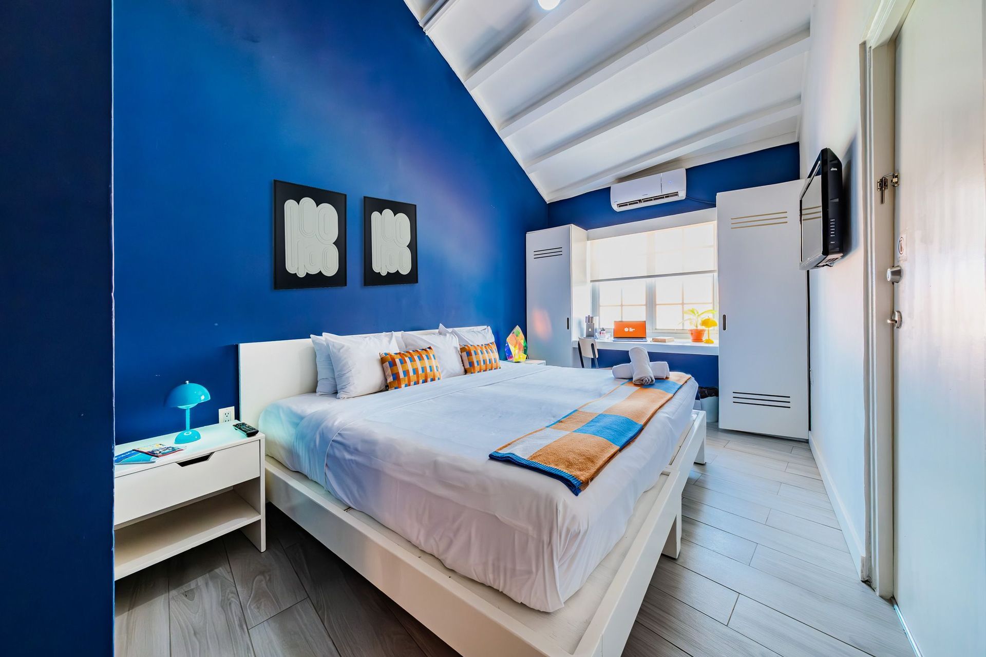 Bright blue bedroom with a double bed, white ceiling beams, and a window letting in daylight