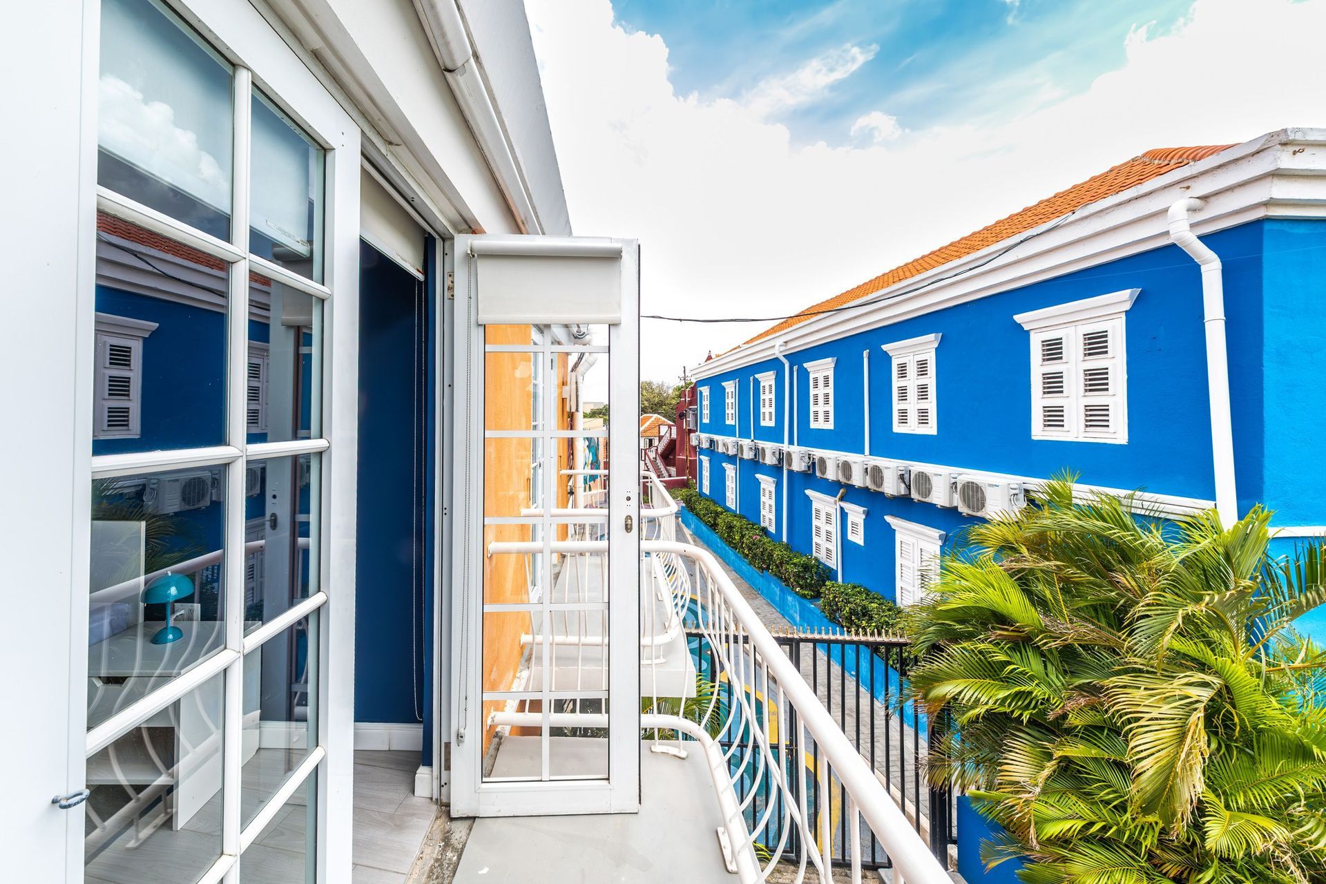 Bright blue building balcony with white railings, open door, and palm tree views under a blue sky