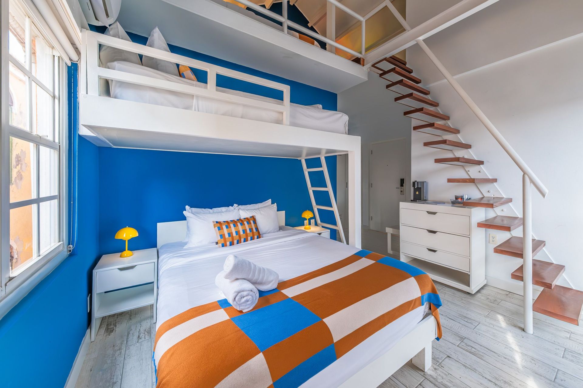 Bright loft bedroom with blue accent wall, bunk bed, staircase, and orange-blue striped bedding