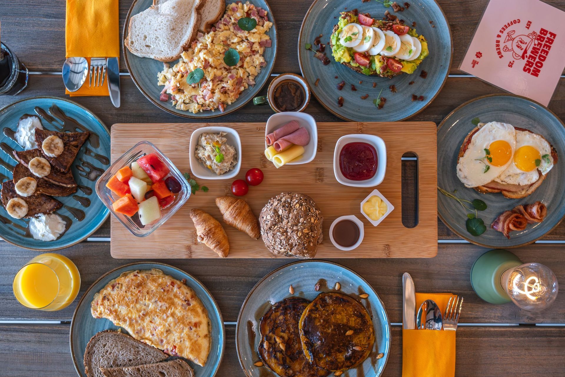 Overhead view of a colorful brunch spread with eggs, toast, fruit, coffee, and sauces on a wooden table