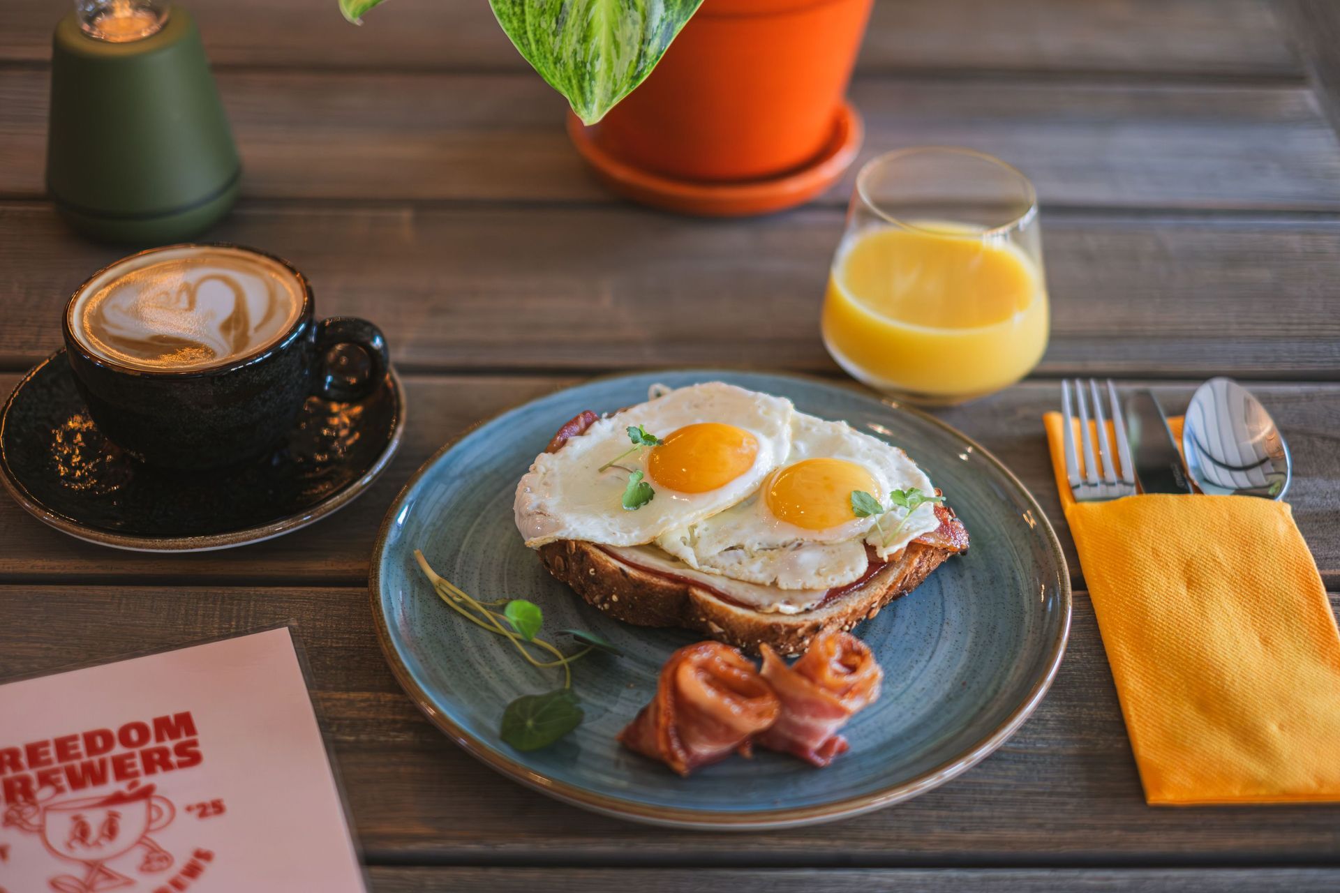 Breakfast on a wooden table: pancakes with fried eggs, bacon, coffee, orange juice, and utensils.