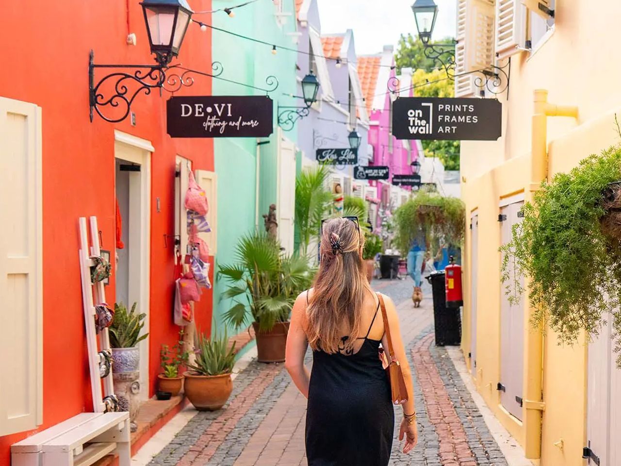 Person walking down a colorful narrow street with pastel buildings, hanging signs, and potted plants