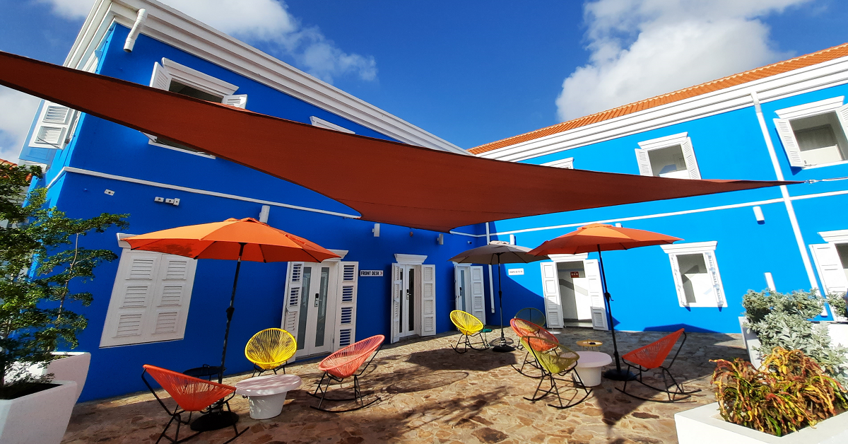 Bright blue courtyard with orange umbrellas, red chairs, and a sunny patio under shade sails.