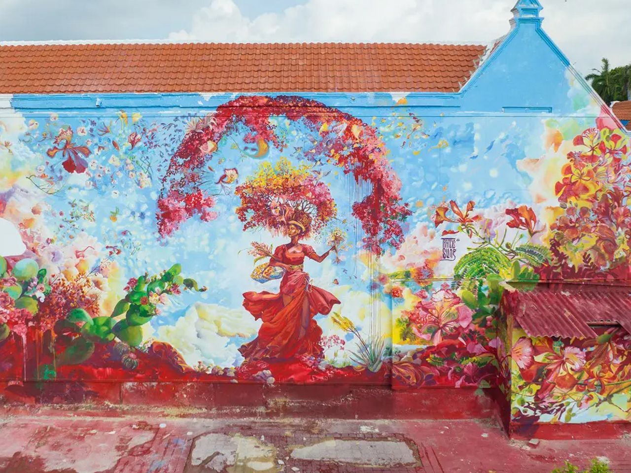 Colorful mural of a red-dressed dancer under a flower arch on a blue wall, with plants and a red-tiled roof