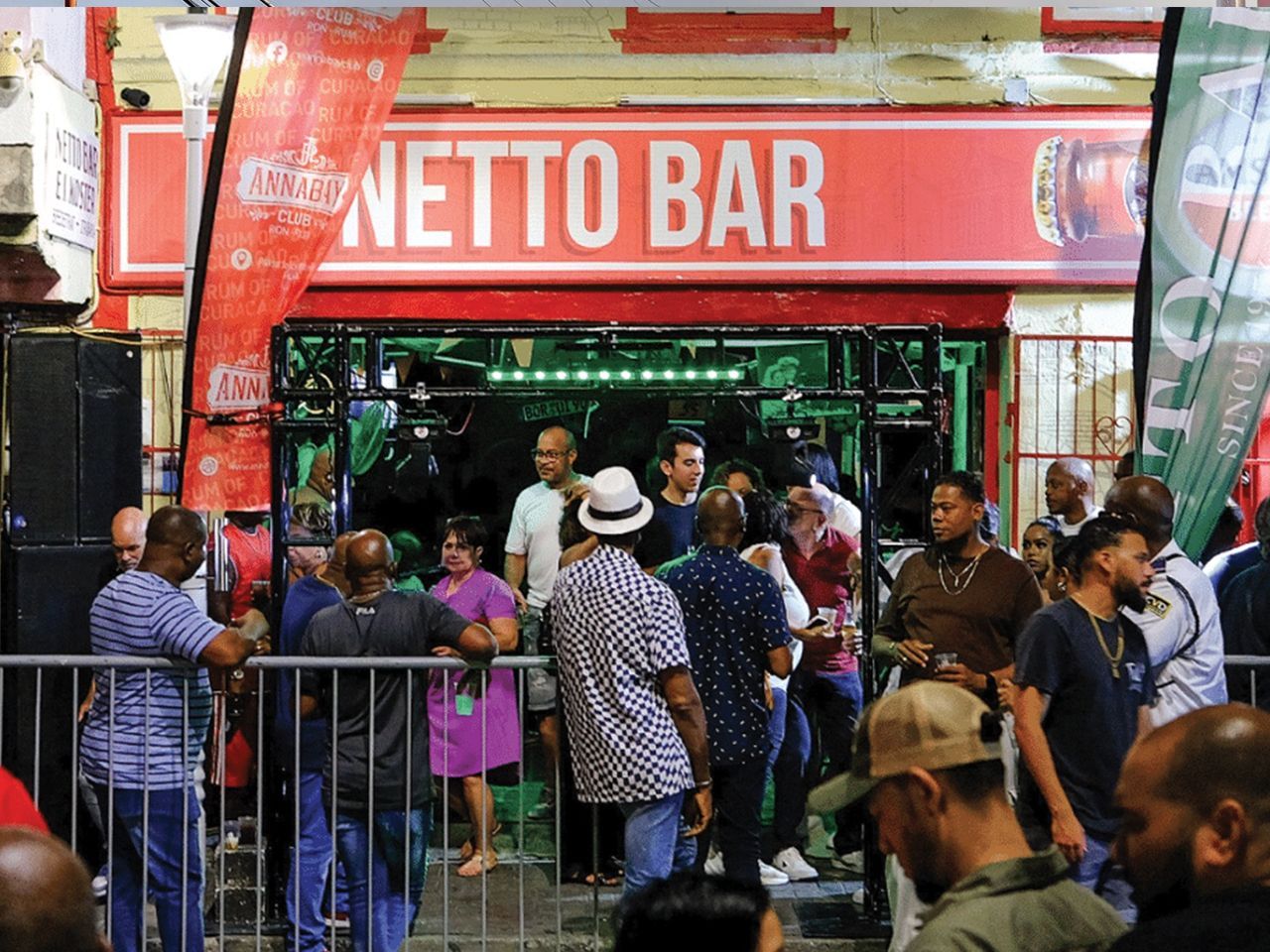Crowded bar entrance with red “NETTO BAR” sign and people gathered outside behind a fence