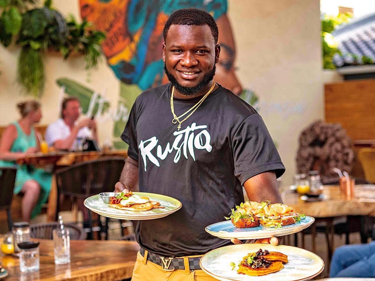 Smiling server in a black “Rugto” shirt carrying three plated dishes in a restaurant