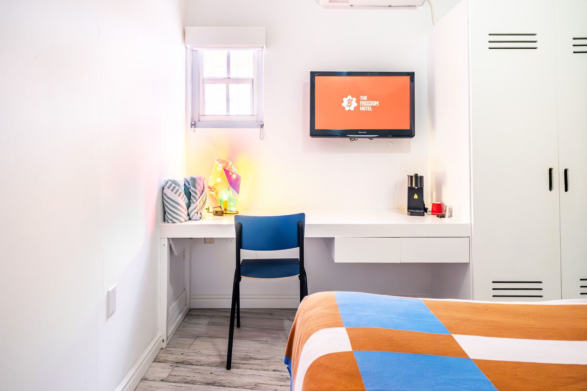 Modern bedroom with white desk, blue chair, wall-mounted TV, and orange-blue bedspread