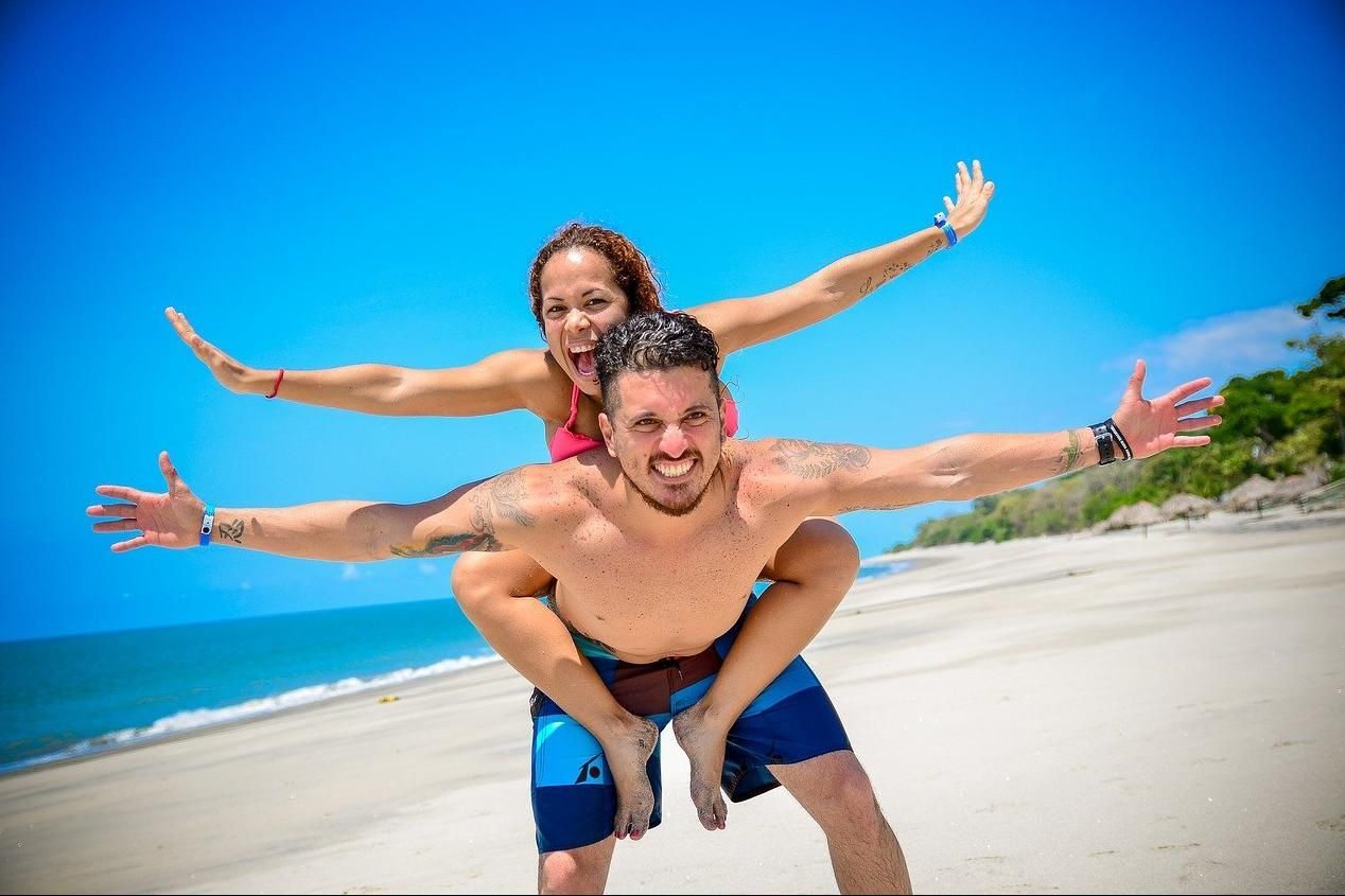 Two people smiling and posing on a sunny beach with arms outstretched near the shoreline