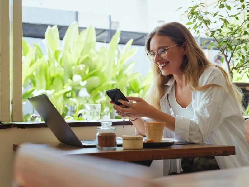 Woman smiling at a café table, using a smartphone beside an open laptop, with bright greenery in the background.