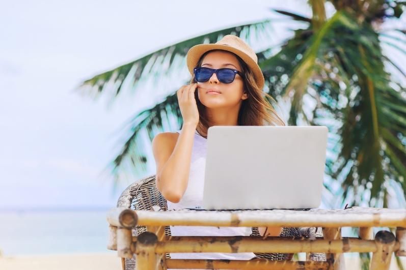 Woman in a straw hat and sunglasses using a laptop at a tropical outdoor table