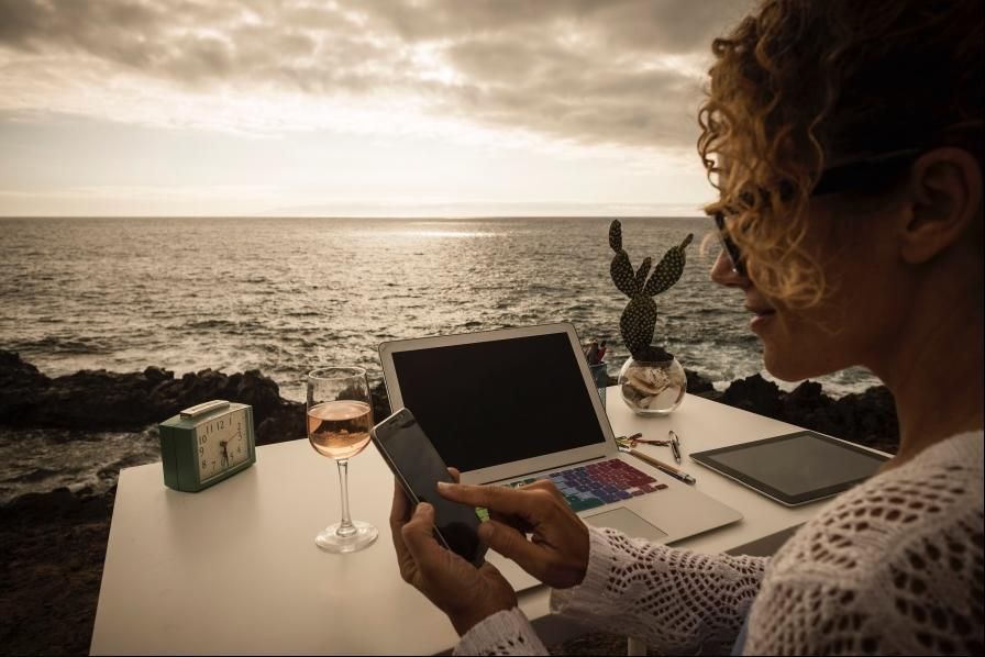 Person using a laptop and phone at a seaside table at sunset, with a wine glass and ocean view.
